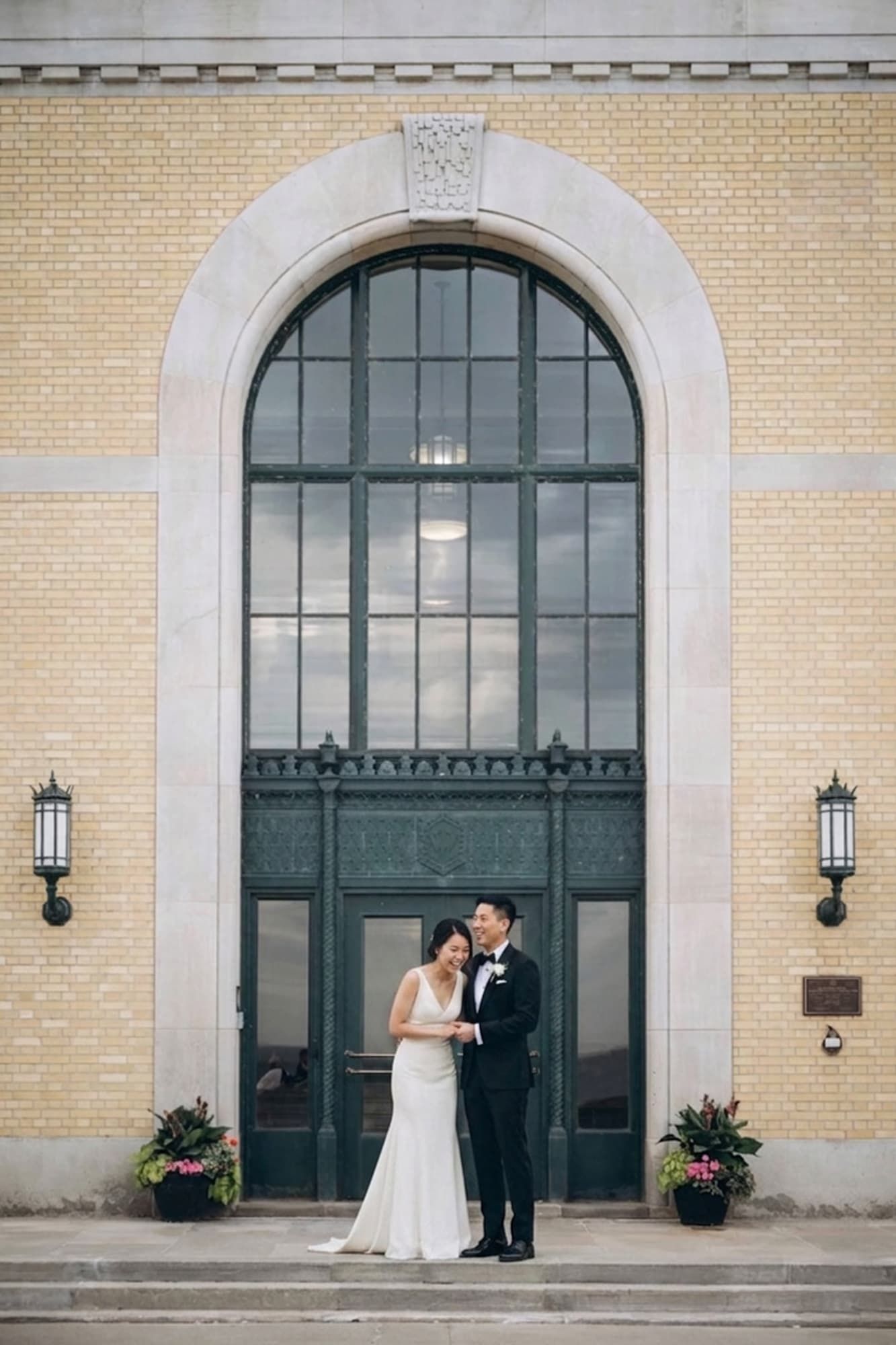 Bride and groom pose before grand arched window of elegant brick building on their wedding day.