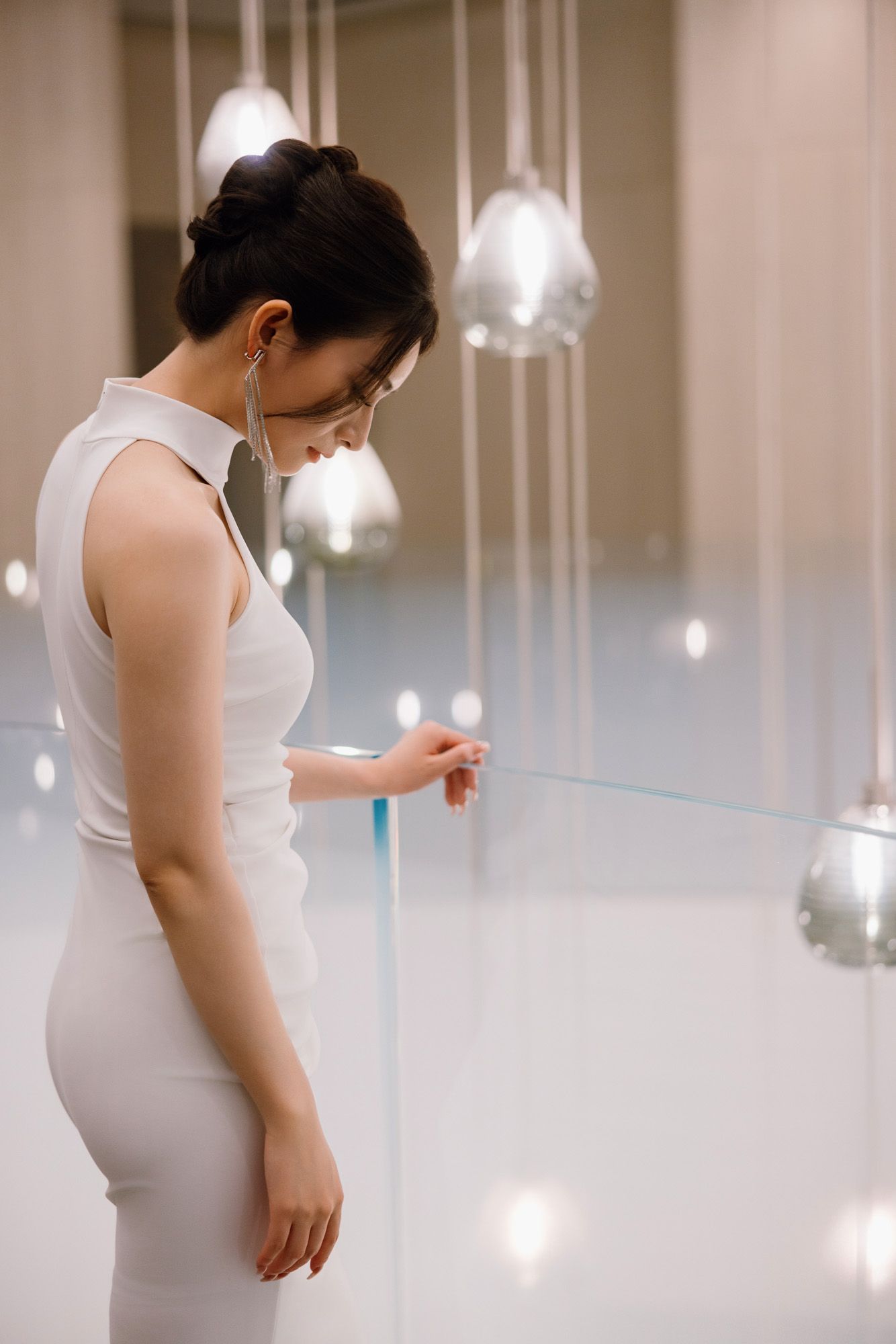 Side profile of bride in white halter dress admiring hanging glass pendant lights at venue.