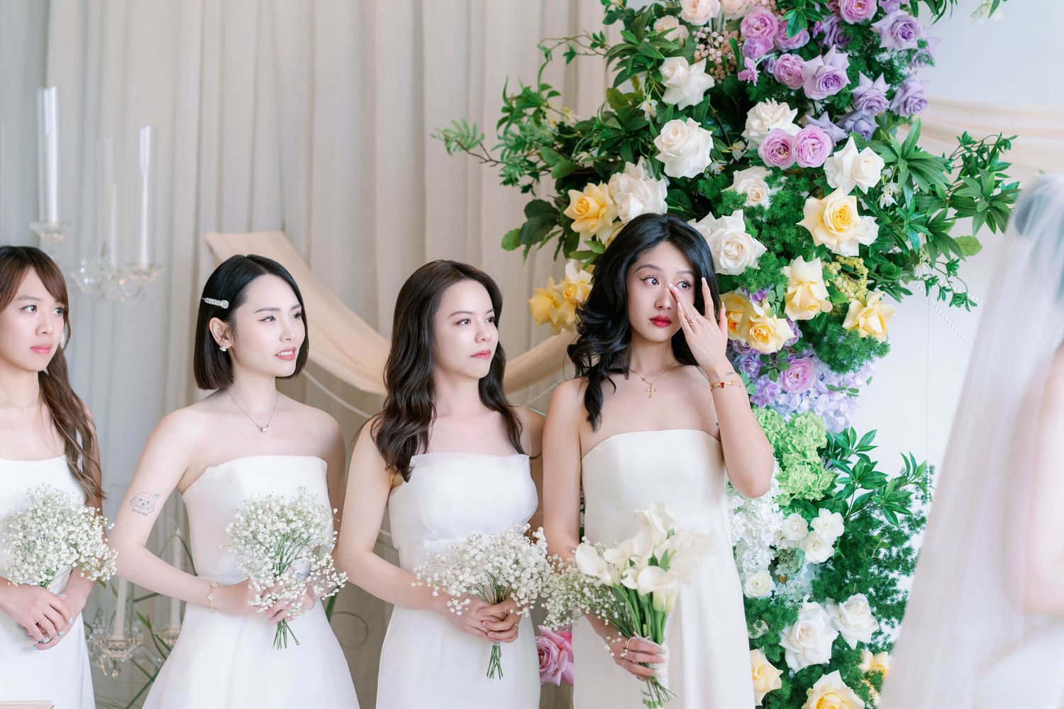Bridesmaids in white strapless dresses holding floral bouquets stand beside colorful flower arch.