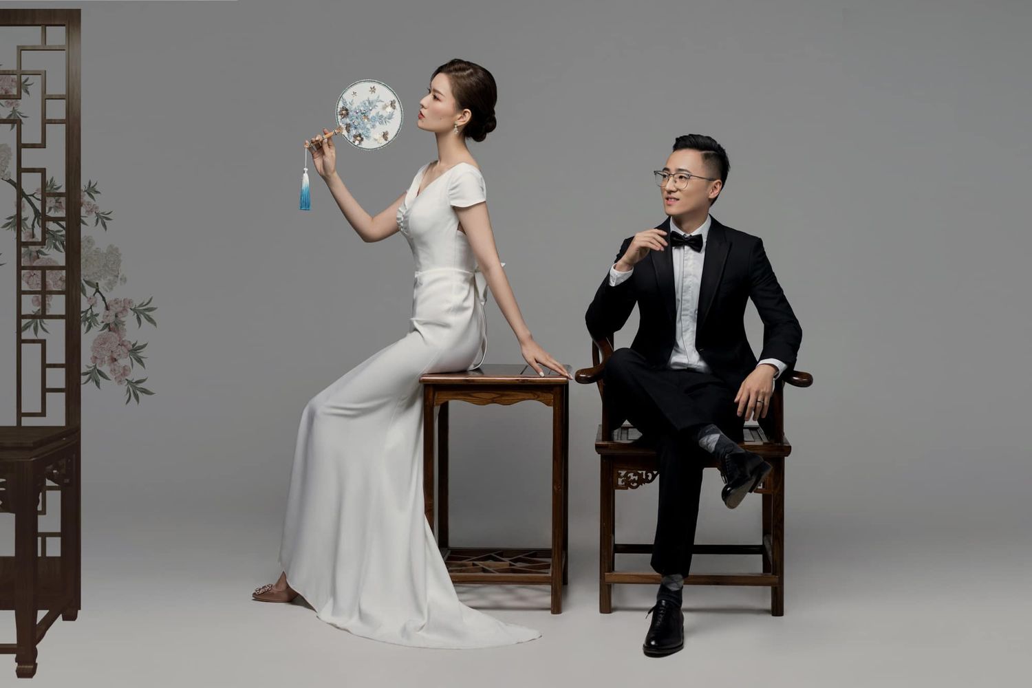 Elegant couple in modern Chinese wedding attire posing with blue bouquet and wooden furniture on grey backdrop.