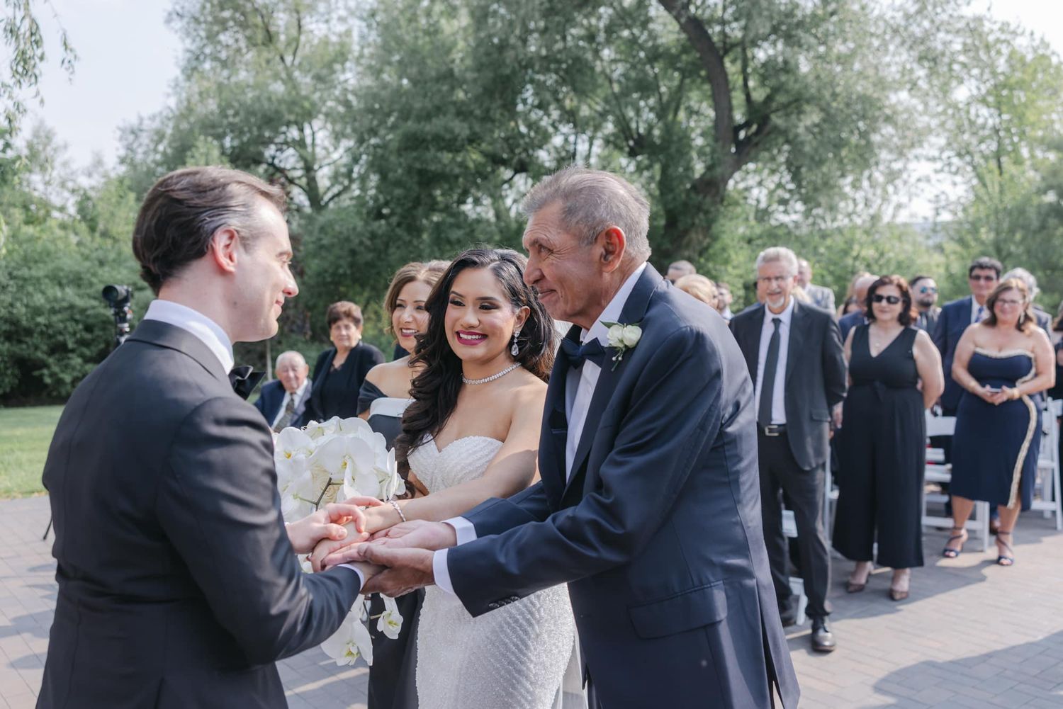 Father of bride presents daughter to emotional groom during outdoor wedding ceremony.