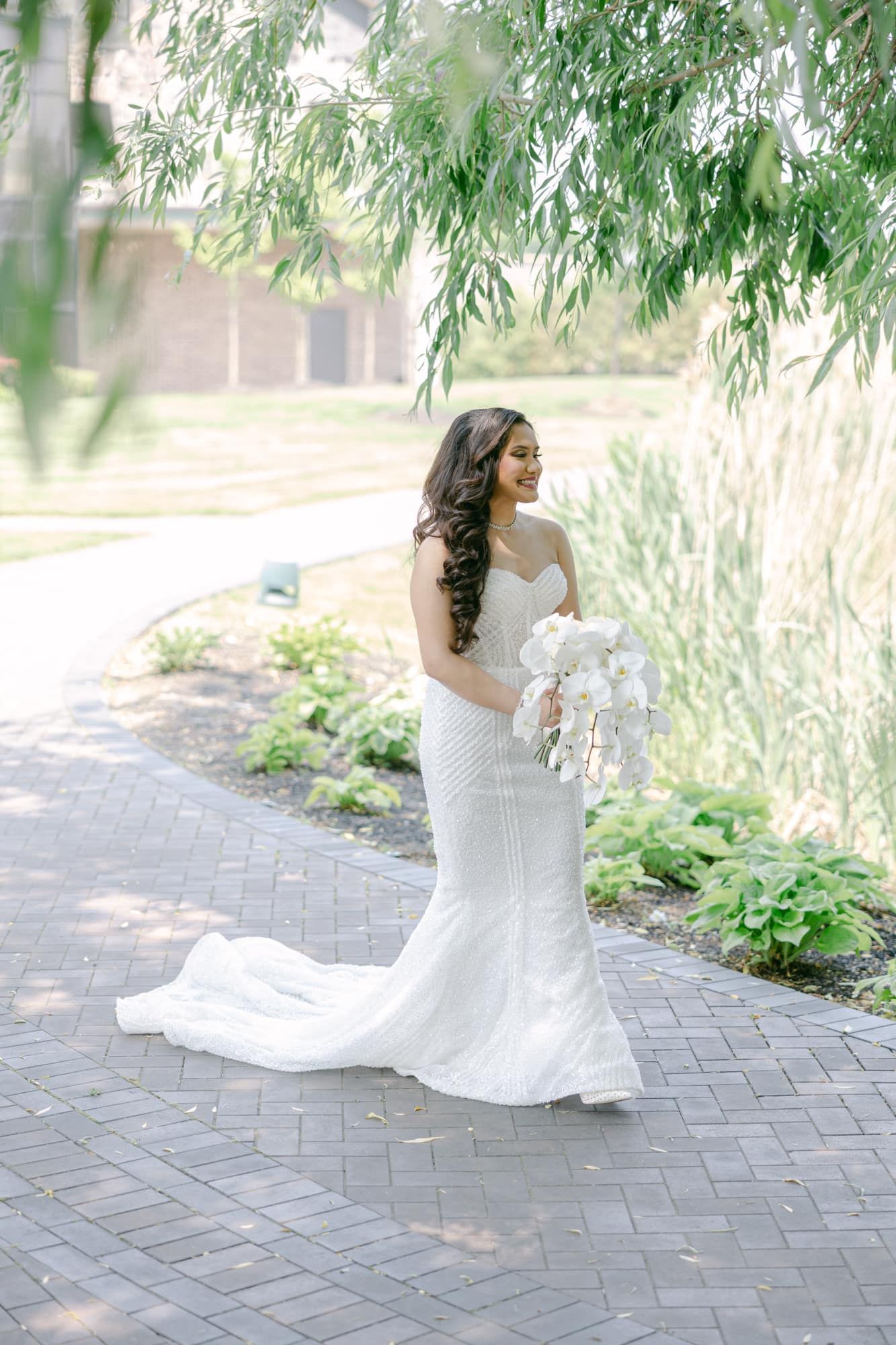 Elegant bride in a fitted white lace gown holding a white bouquet while walking under willow trees.