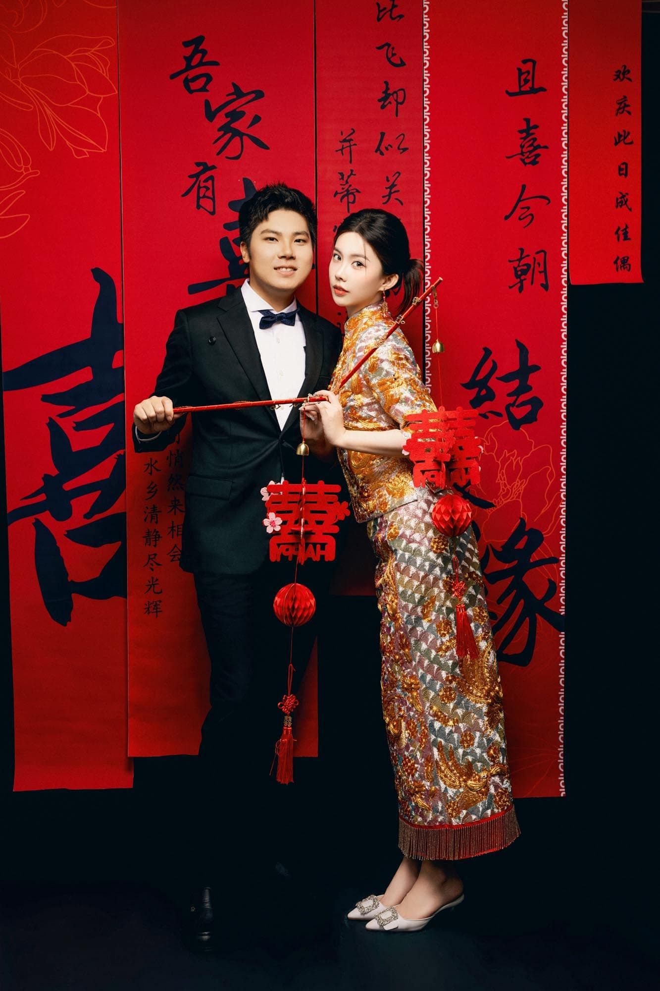 A couple poses in traditional Chinese wedding attire before red banners with calligraphy, holding decorative red accessories.