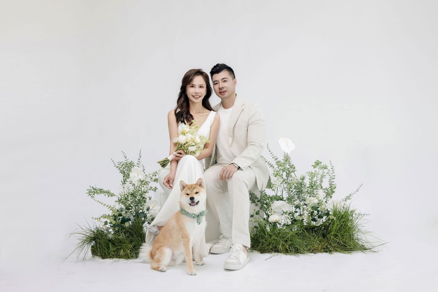 Couple in white outfits posing with a Shiba Inu dog among green plants and white flowers on a white background.