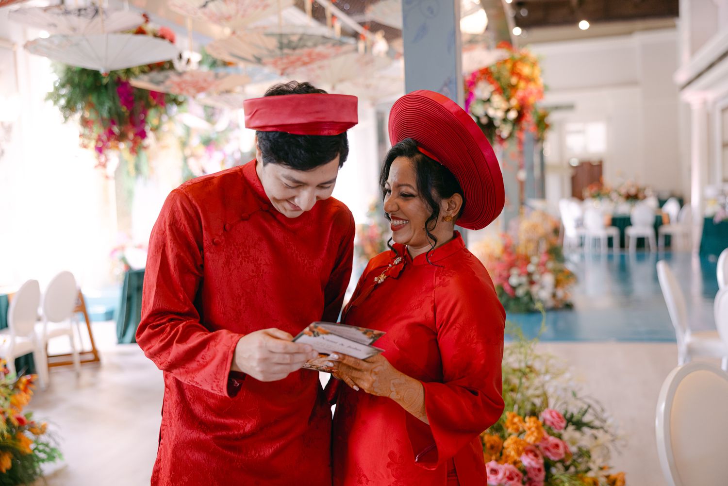 A couple in matching red traditional Vietnamese wedding attire read a card together amid colorful floral decorations.