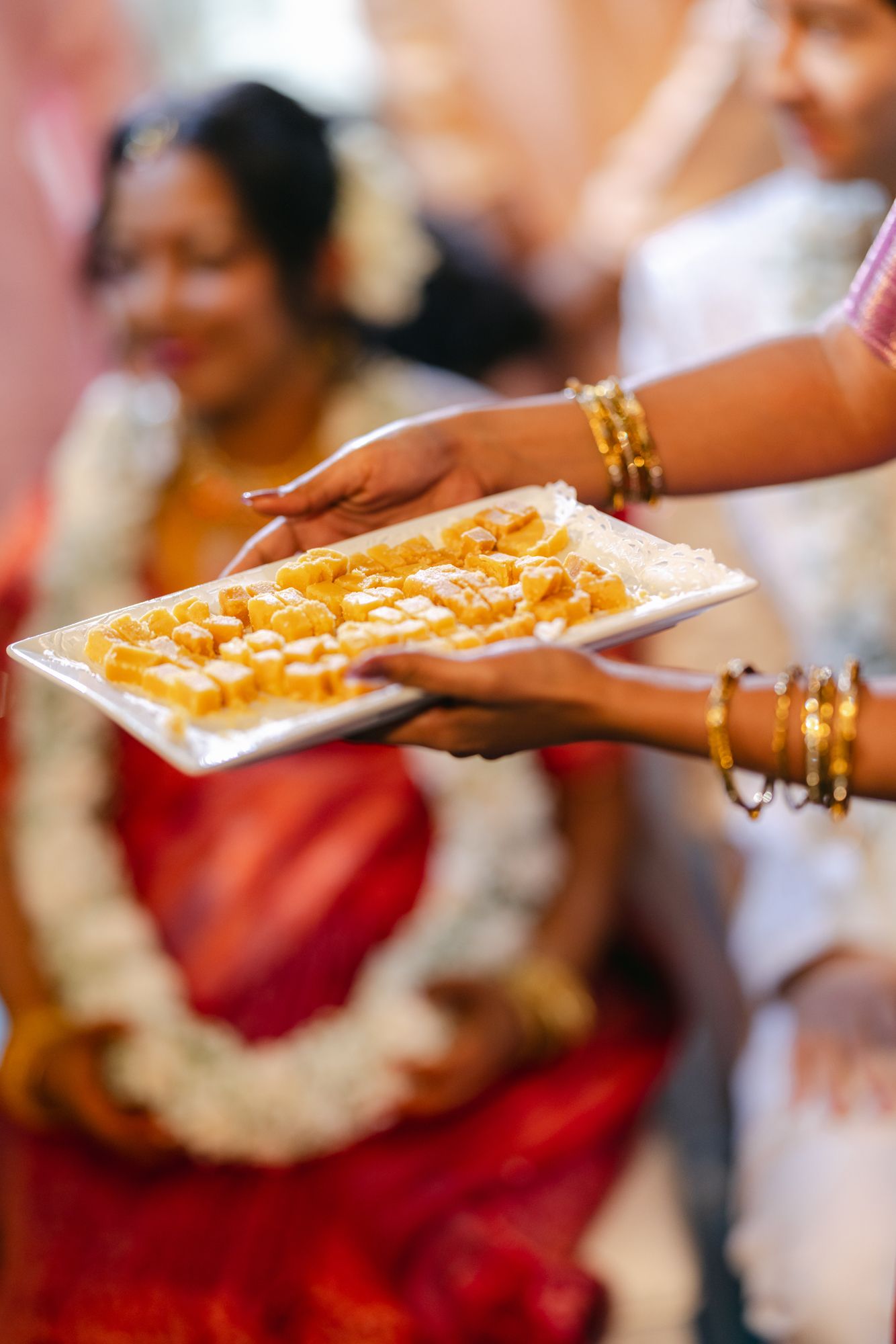 Hands holding a white tray of golden Indian sweets during a wedding ceremony, with a woman in a red sari and garland behind.
