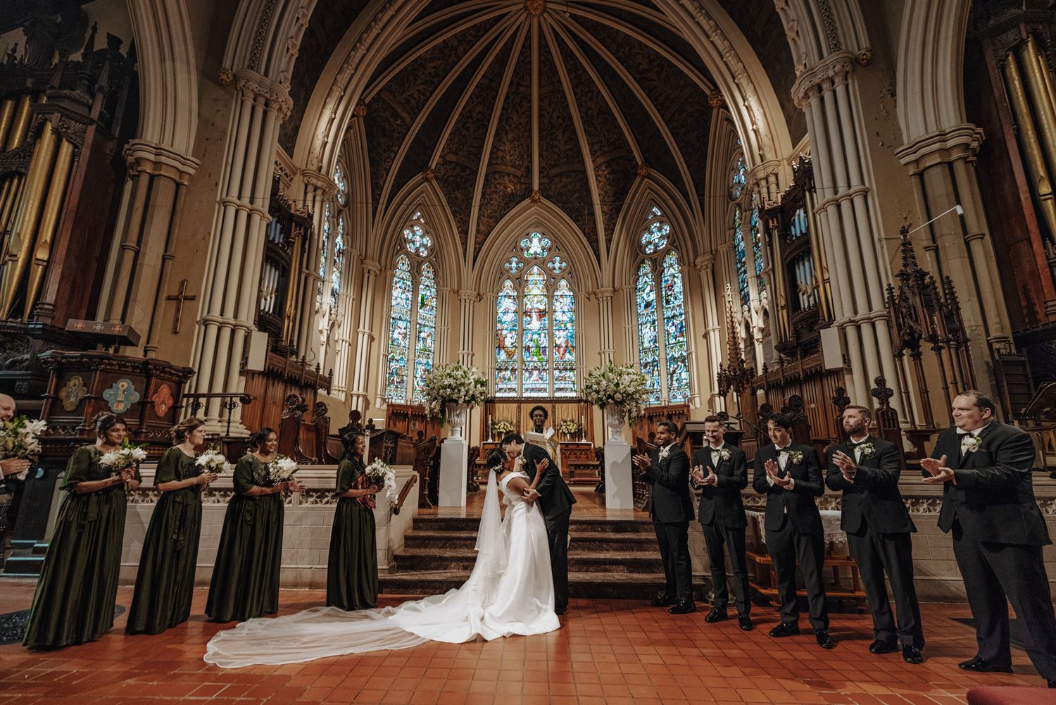 Bride and groom stand at the altar of a historic Gothic church with bridesmaids and groomsmen lined up on both sides.
