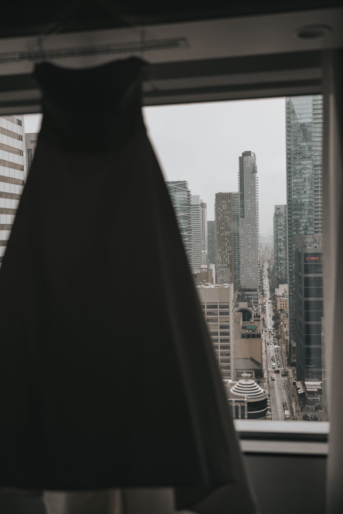 Strapless wedding dress hanging in a hotel window silhouetted against a misty city skyline of high-rise buildings.