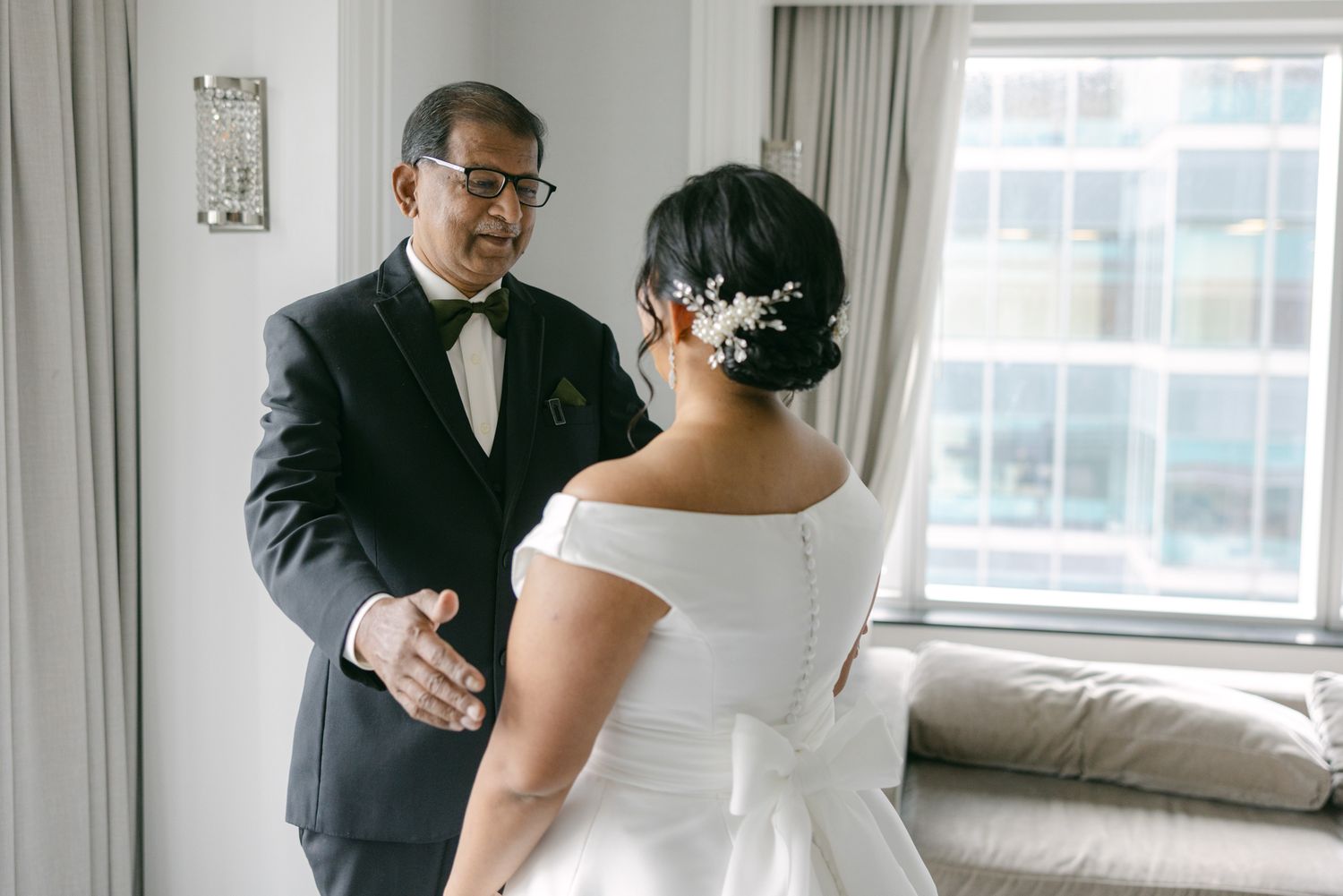 Father in a black tuxedo and bow tie gets an emotional first look at his daughter, a bride in a white gown with floral hair accessory.
