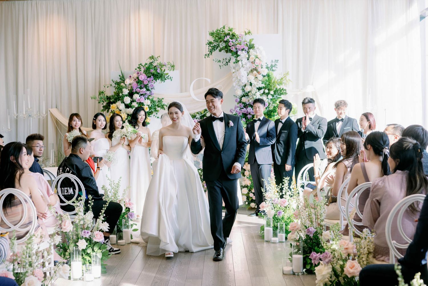 Bride and groom walk triumphantly down aisle past seated guests after indoor wedding ceremony.