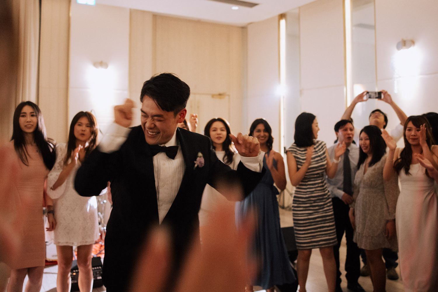 Groom in tuxedo dances energetically with fist pump as wedding guests cheer and photograph the moment.