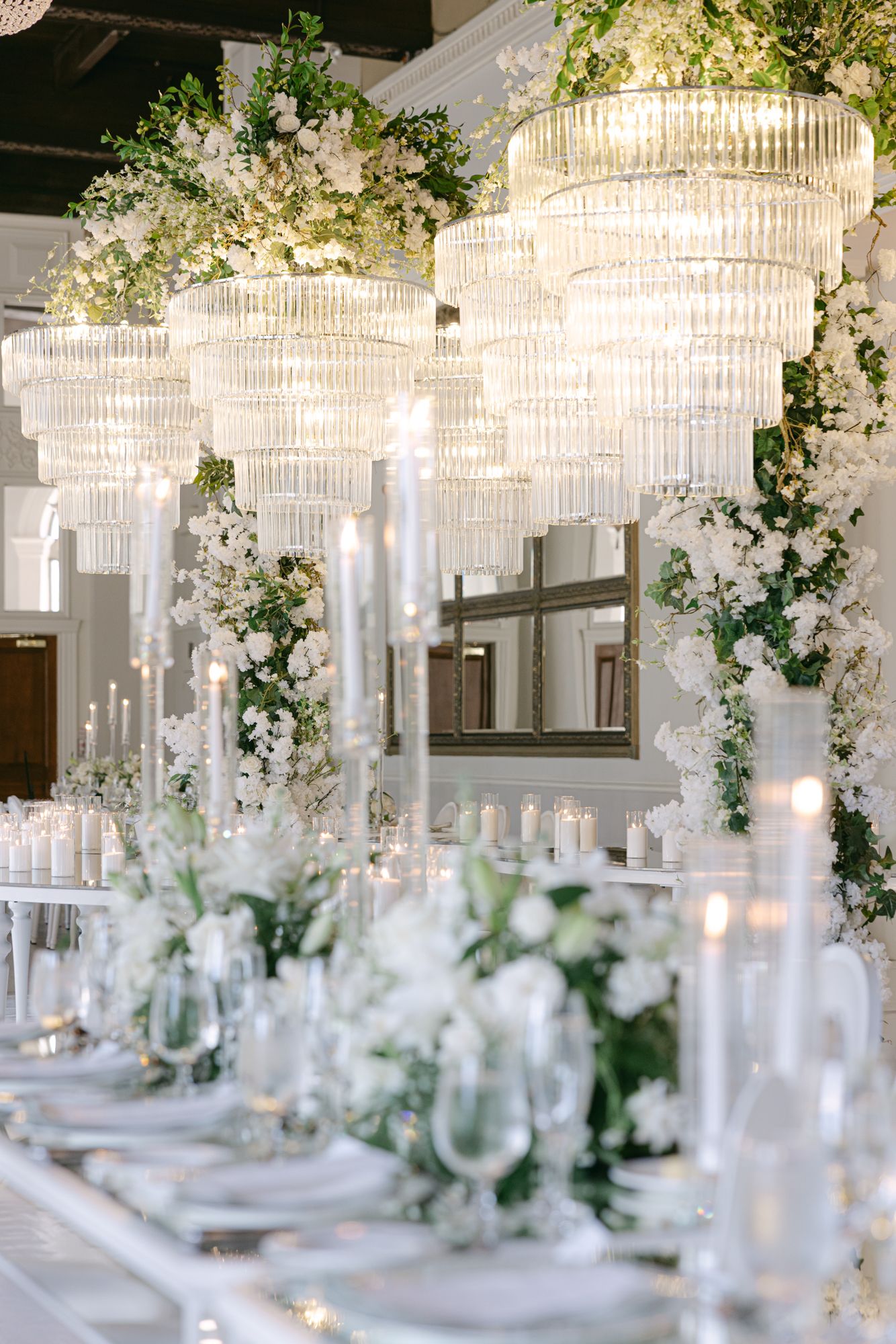 Elegant wedding reception table with white floral centerpieces, tall taper candles, and tiered crystal chandeliers draped with greenery.