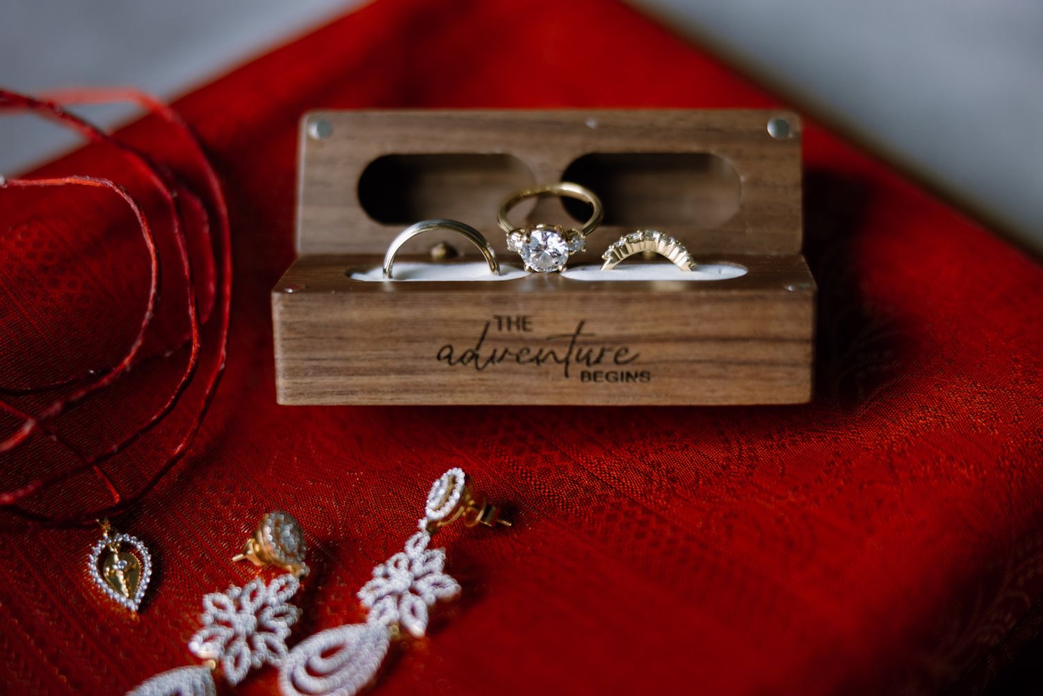 Wooden ring box holding engagement and wedding rings on a red cloth, beside dangling bridal earrings and a leaf pendant necklace.