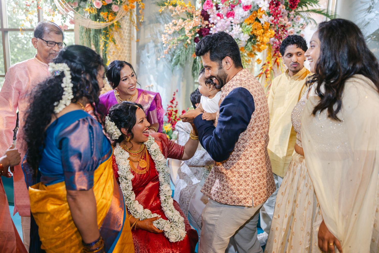 Family and guests in colorful traditional attire gather around a bride in a red sari and floral garlands at a Hindu wedding.