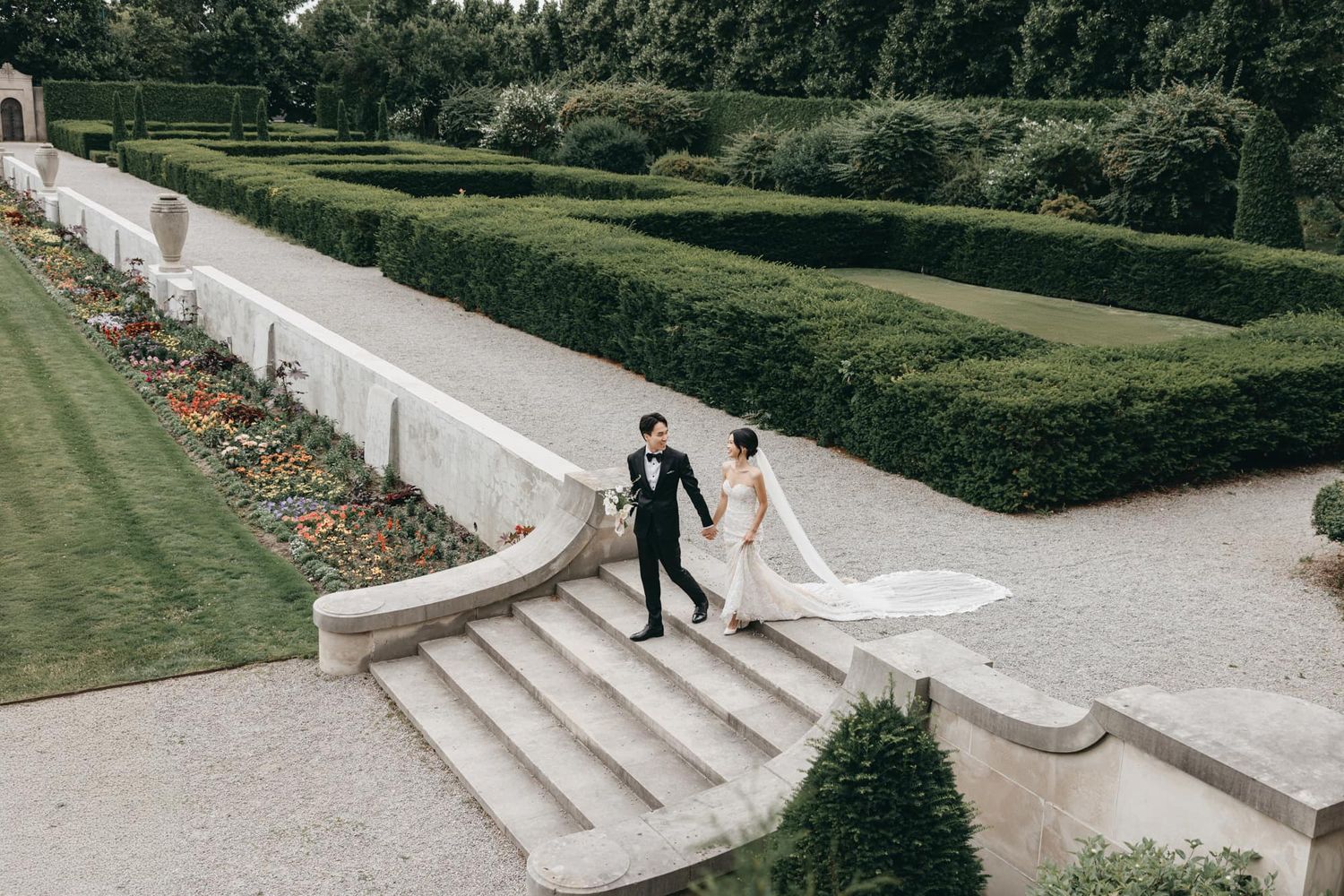 Bride and groom walking hand in hand down stone steps in a formal garden with manicured hedges.