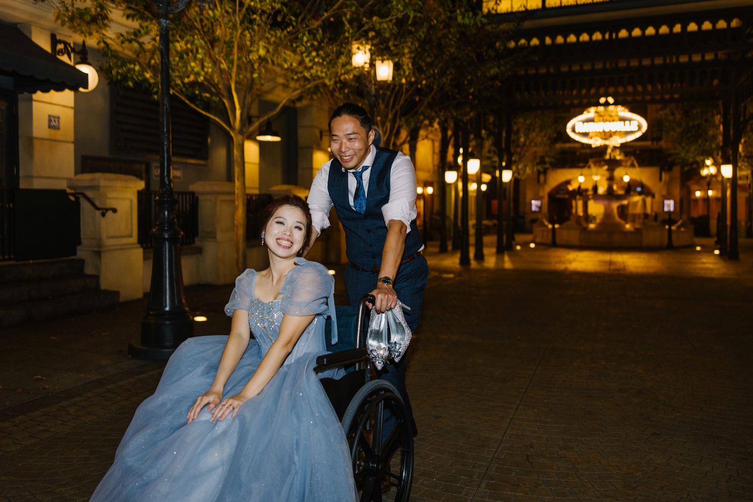 Joyful couple in blue evening wear posing with a vintage bicycle on a charming night-lit street.