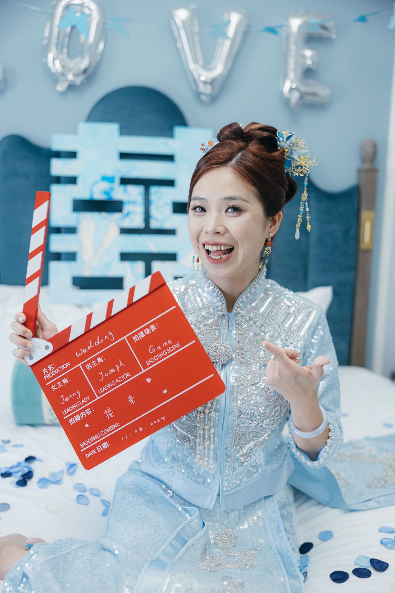 Bride in blue qipao holding red film clapperboard on decorated bed with LOVE balloons.