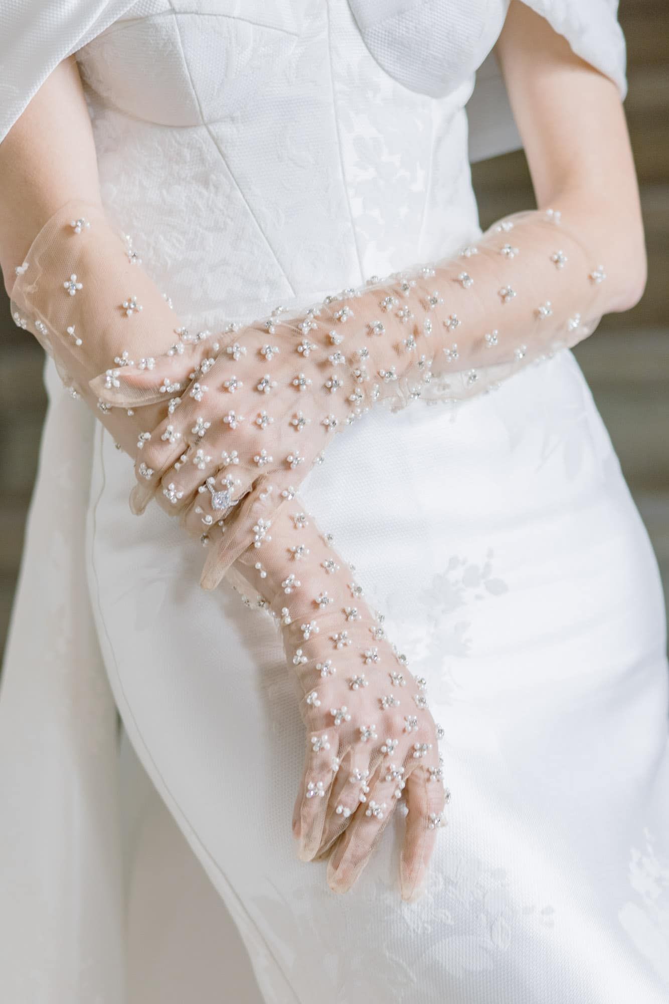 Bride wears elegant sheer tulle gloves adorned with pearl and floral embellishments on wedding day.