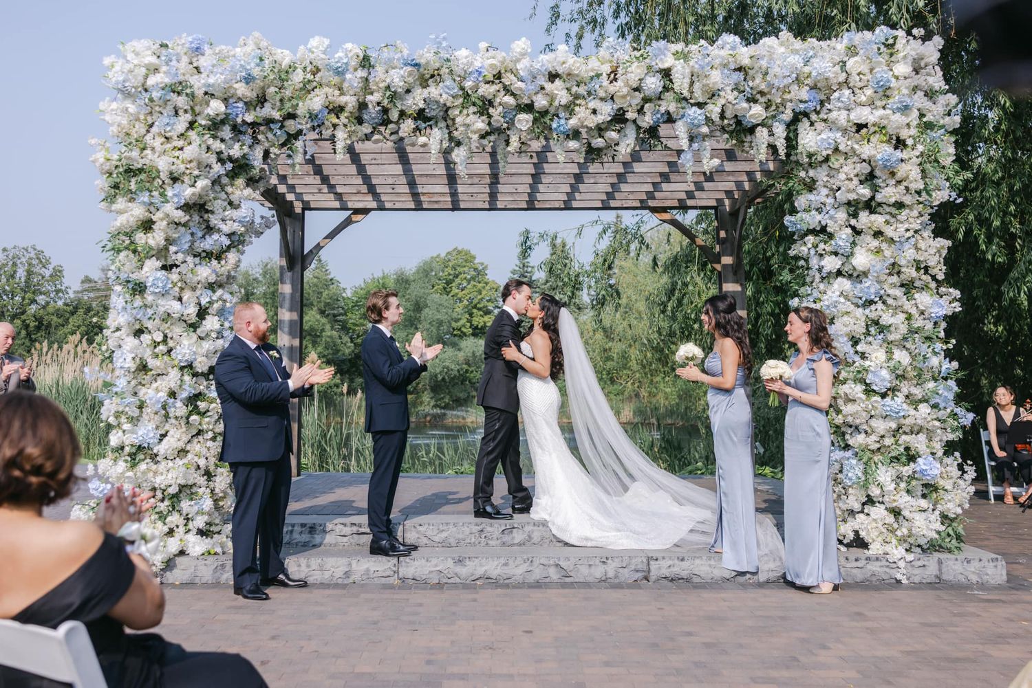 Newlyweds share first kiss under stunning floral arch at outdoor wedding ceremony.