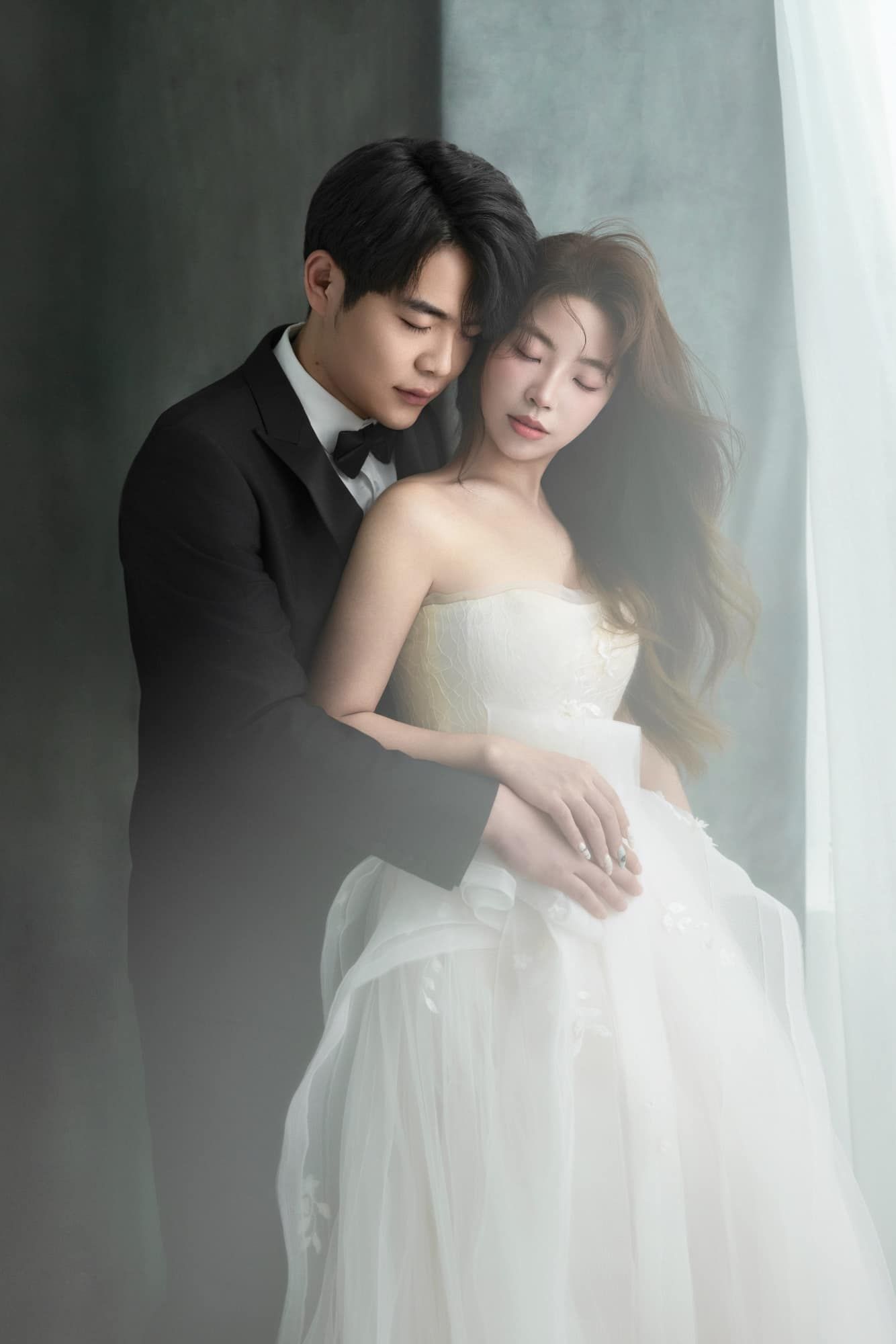 Romantic Asian couple in black tuxedo and white strapless gown embracing by sheer curtains.