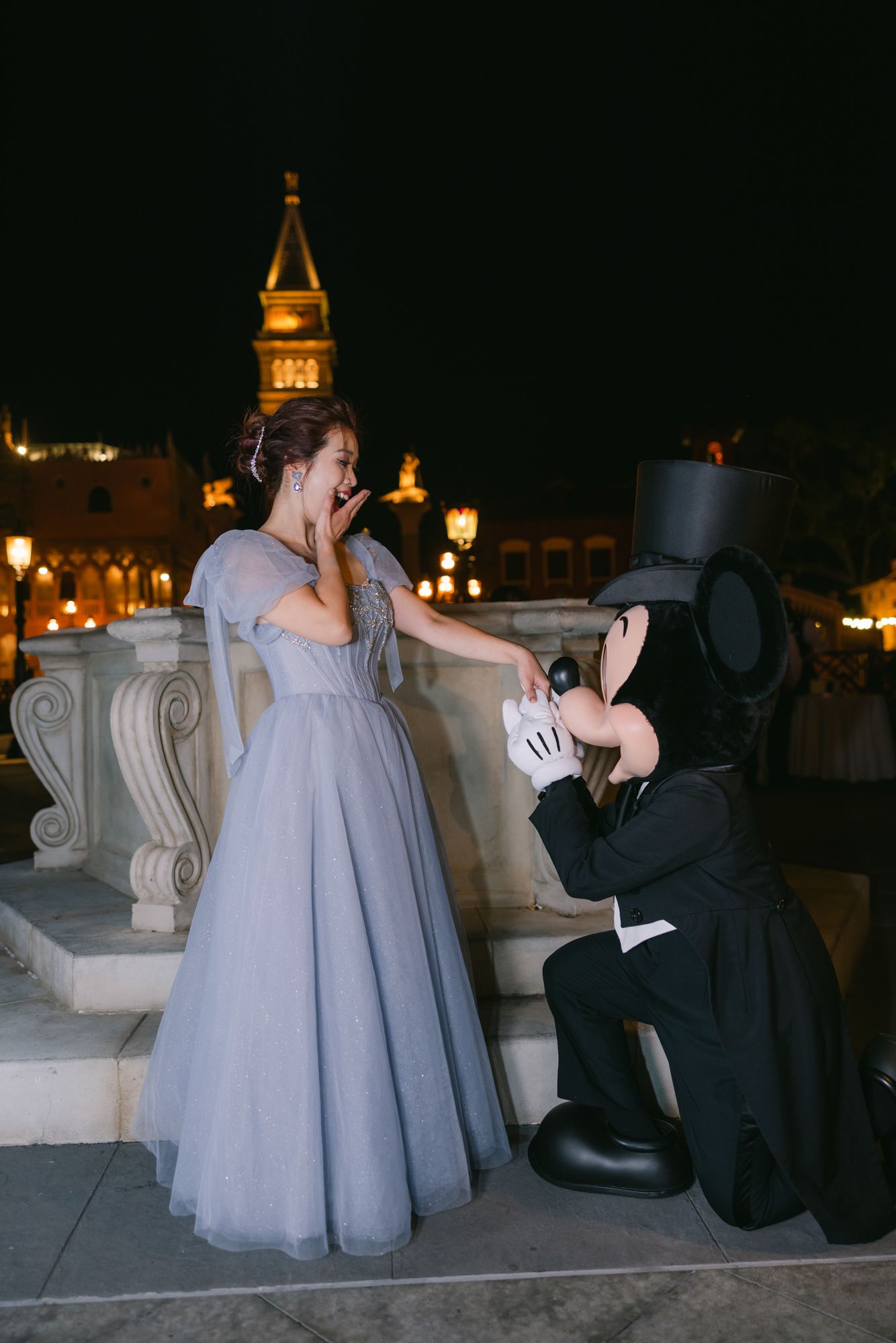 Mickey Mouse kneels before a surprised woman in a blue gown during a magical Disney proposal at night.