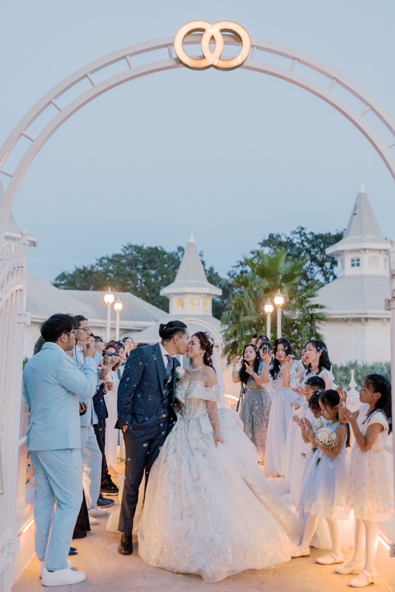 Bride and groom kissing under decorative arch with wedding rings while guests celebrate with confetti.