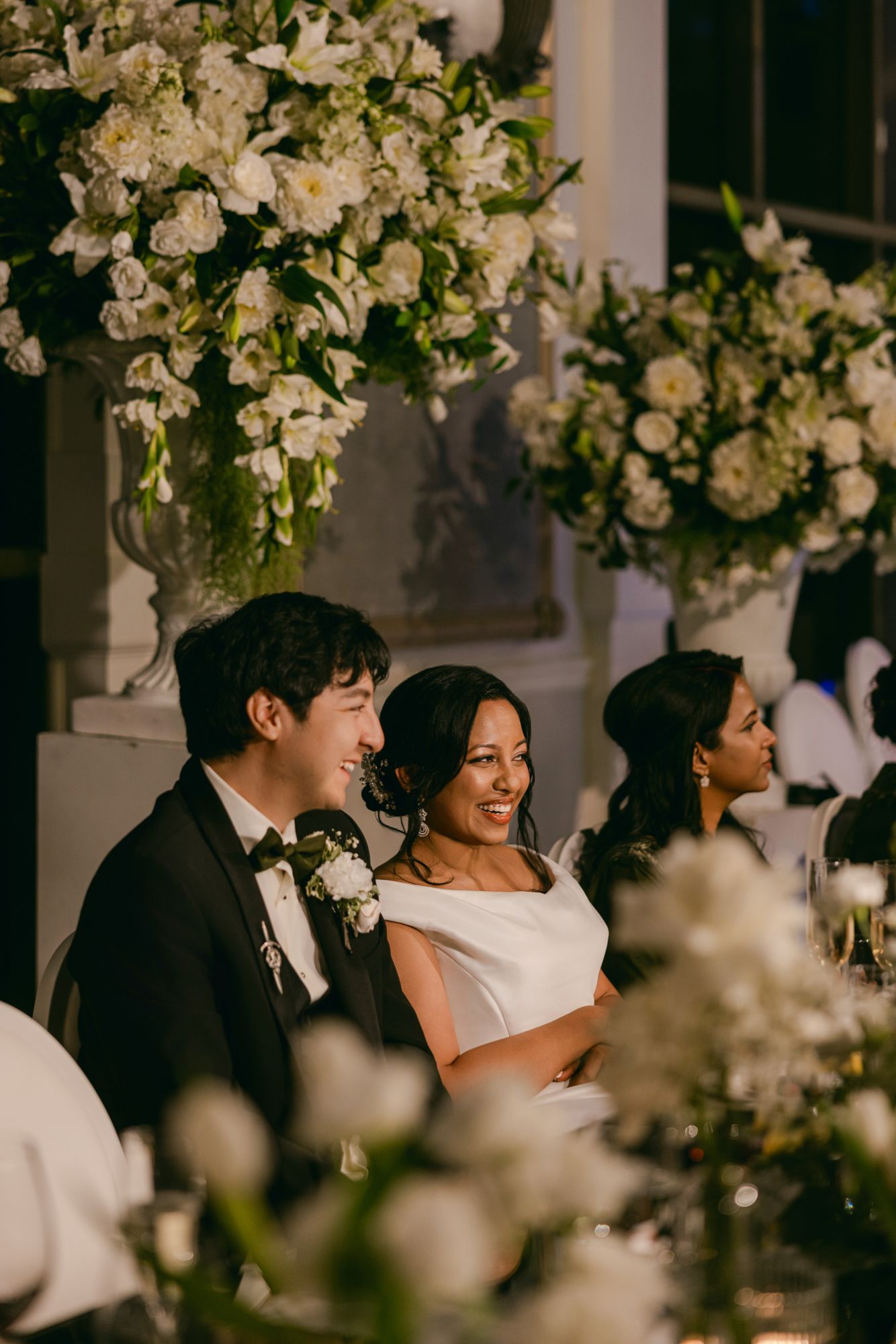 Smiling bride and groom sit at the head table surrounded by lush white floral arrangements during their wedding reception.