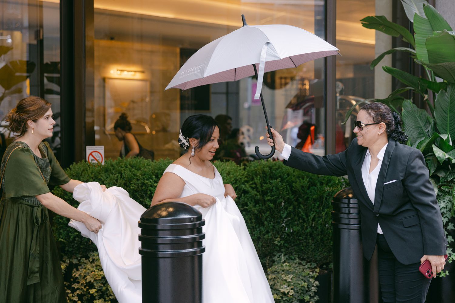 Bride in a white wedding gown walks under a white umbrella held by a guest while a bridesmaid in green helps with her train.