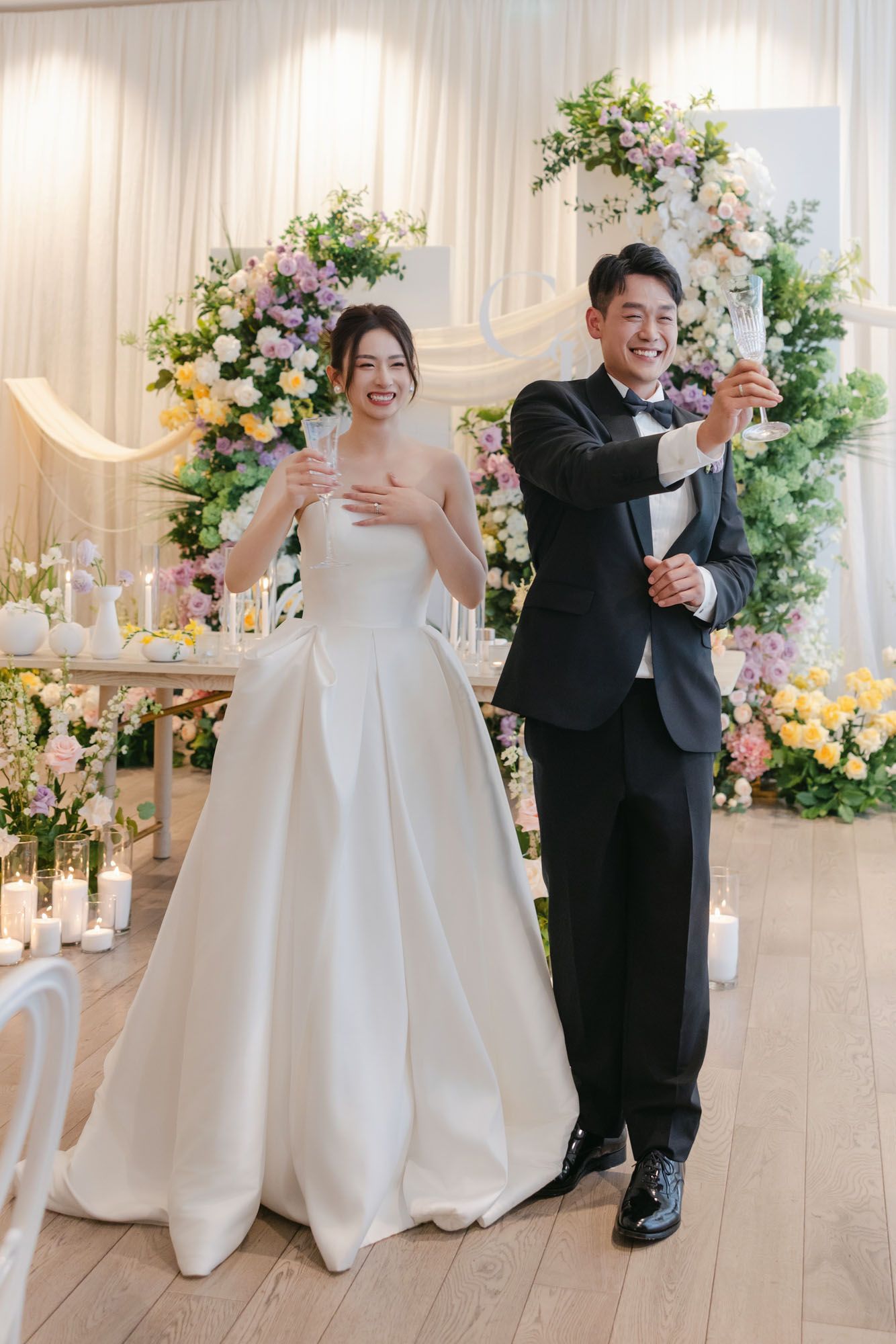 Smiling bride and groom toasting with champagne in front of a colorful floral arch at their wedding.