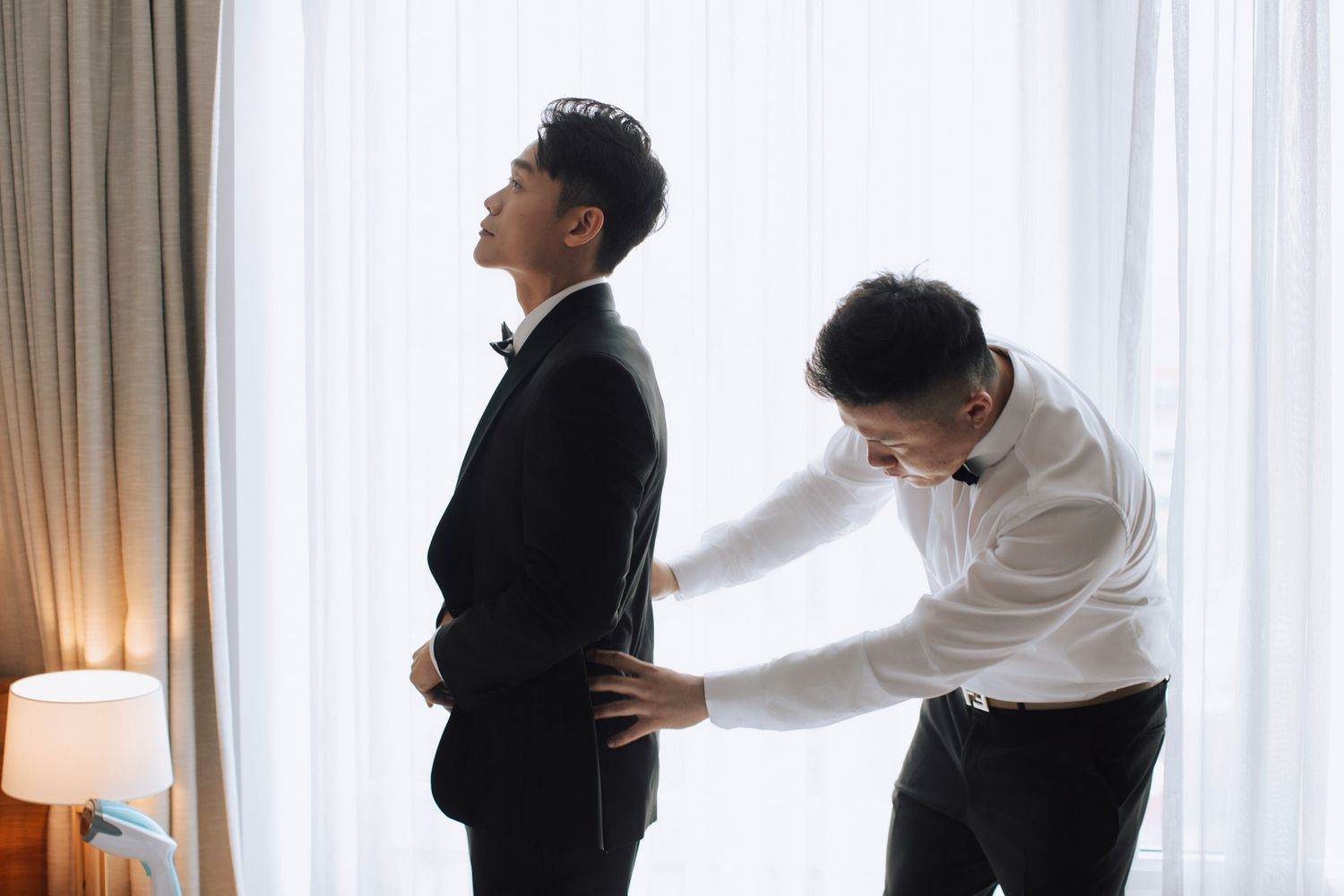 Groomsman helping groom adjust black tuxedo jacket near bright window before wedding ceremony.