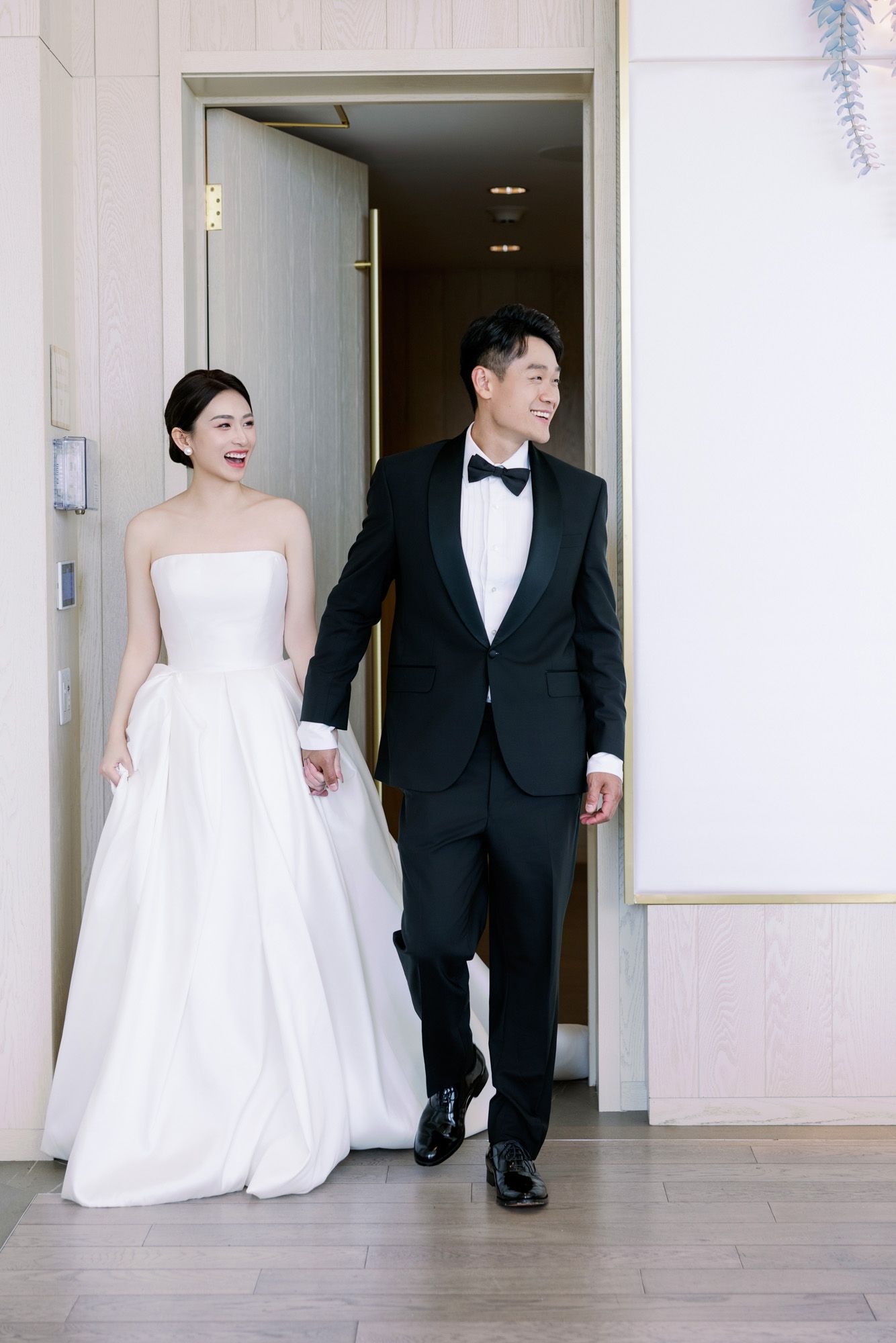Smiling bride and groom in elegant wedding attire walk hand in hand through venue doorway.