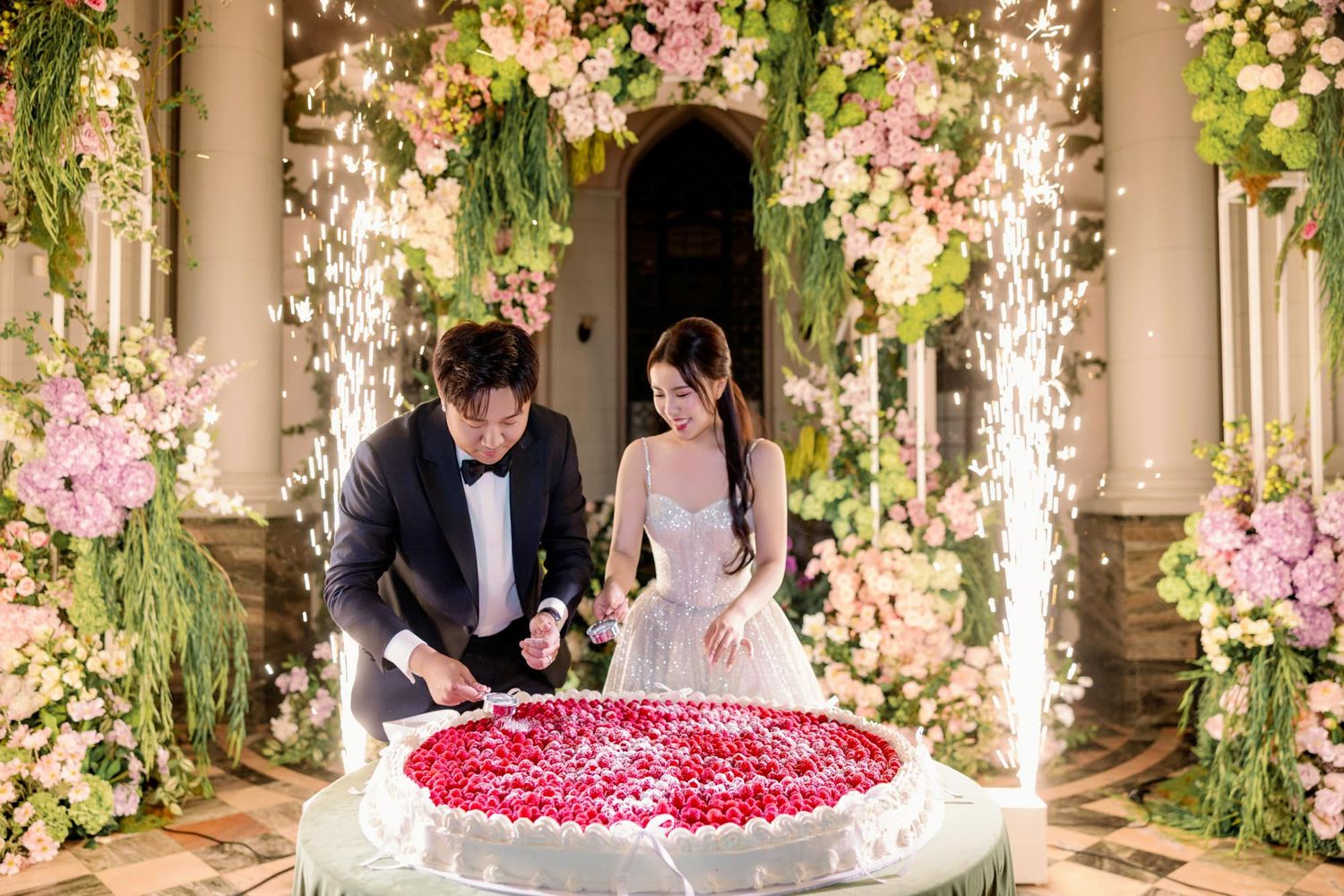 Couple cutting a floral wedding cake surrounded by sparklers and pink flower arrangements.