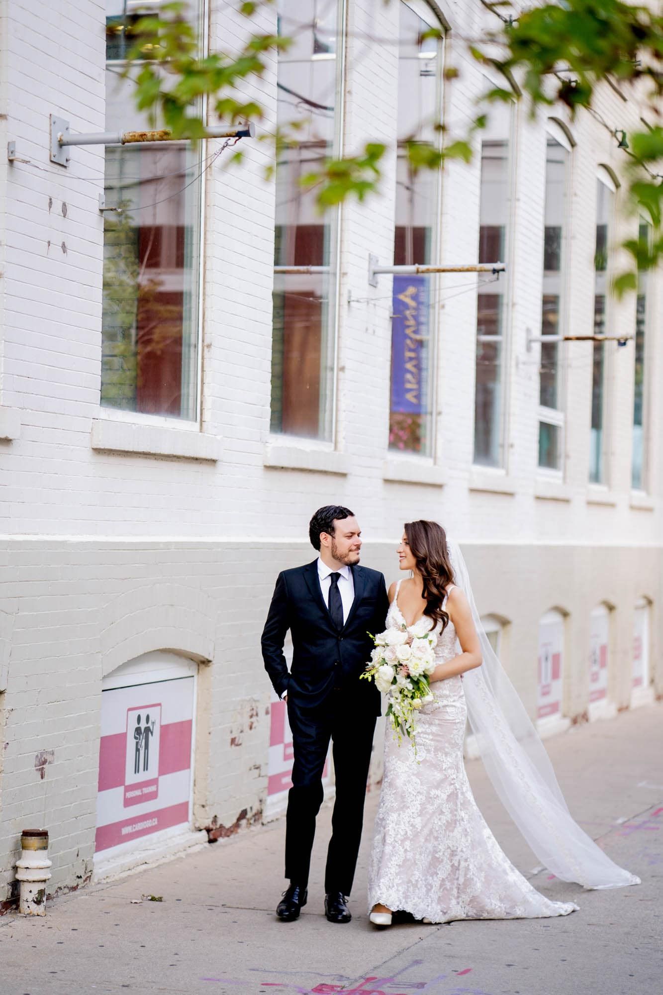 Bride and groom share loving glance while walking on city sidewalk with white bouquet and veil.