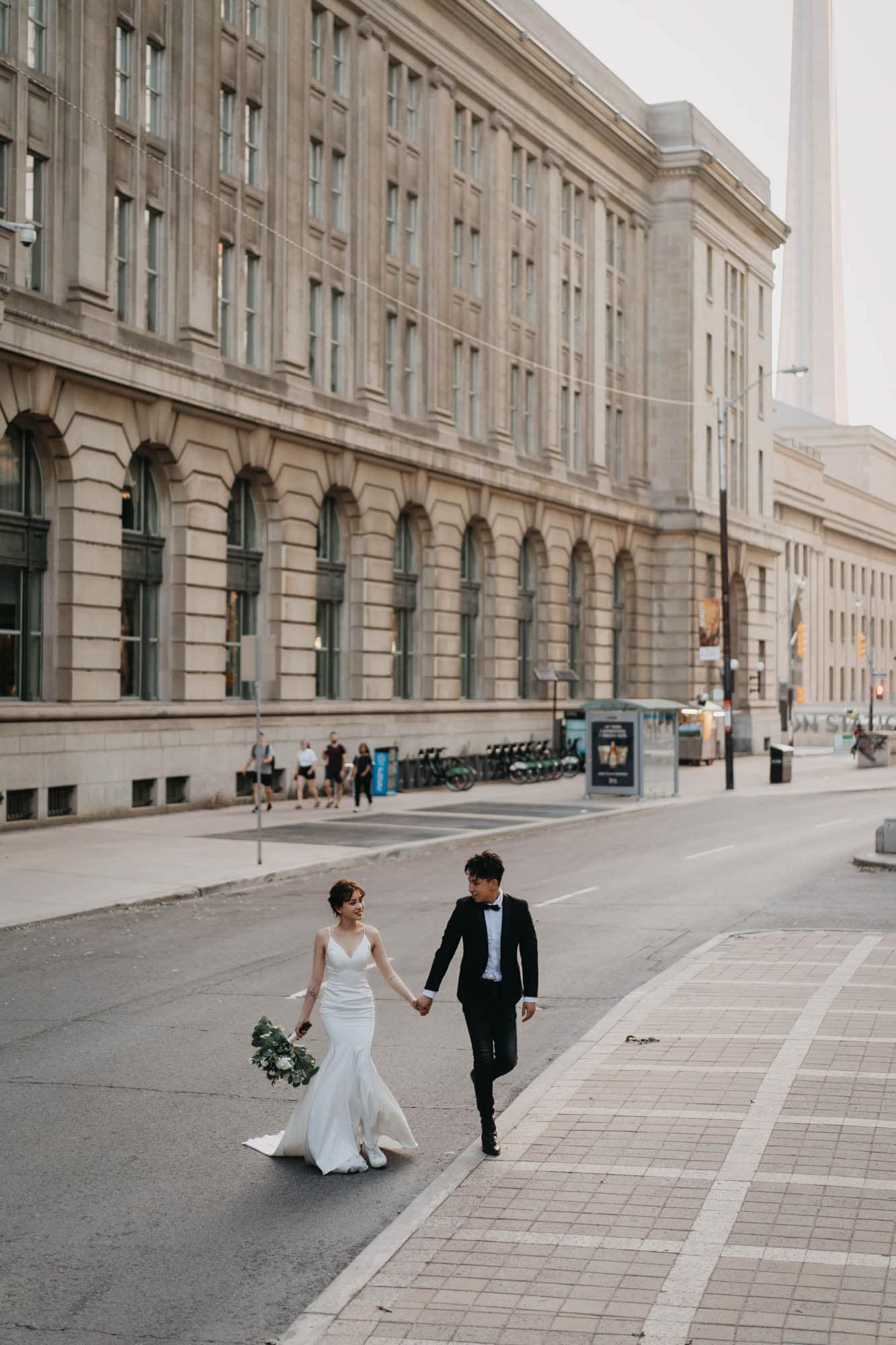 Bride and groom walk hand-in-hand along a grand urban street, with a classic stone building backdrop.