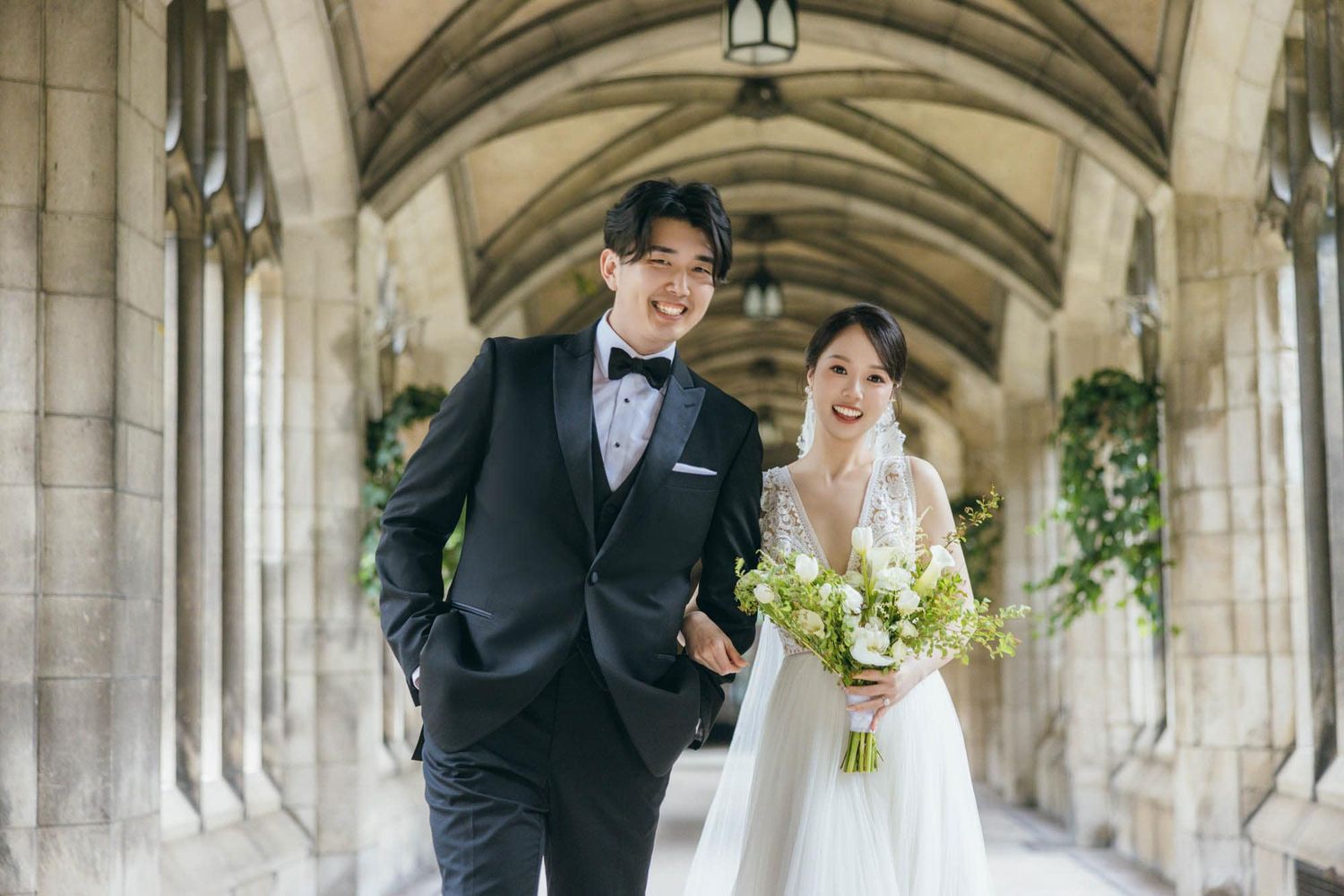 Happy couple in wedding attire walks hand-in-hand under stone arches, bride holding white bouquet.