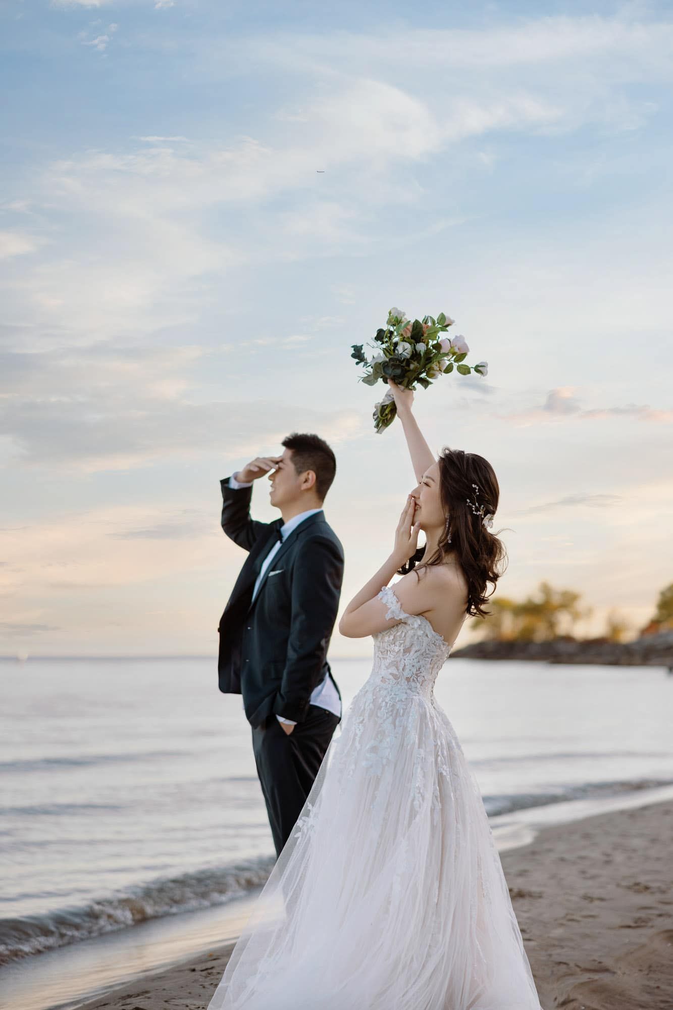 Bride raises bouquet on beach at sunset while groom stands beside her in black suit during romantic seaside wedding.