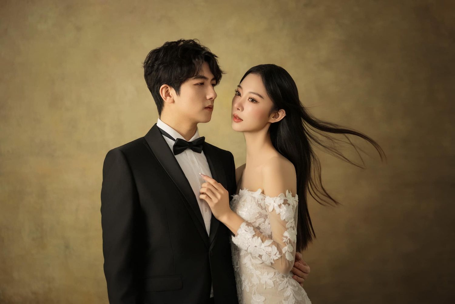 Elegant couple in wedding attire pose for a studio portrait, groom in tuxedo and bride in white off-shoulder gown.