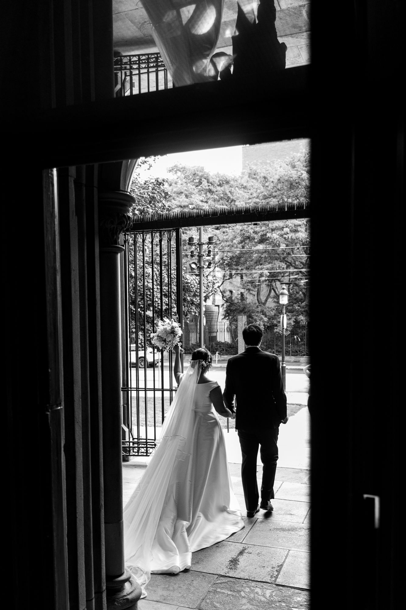 Black and white photo of a bride in a long veil and gown walking arm in arm with her groom out of a church doorway.