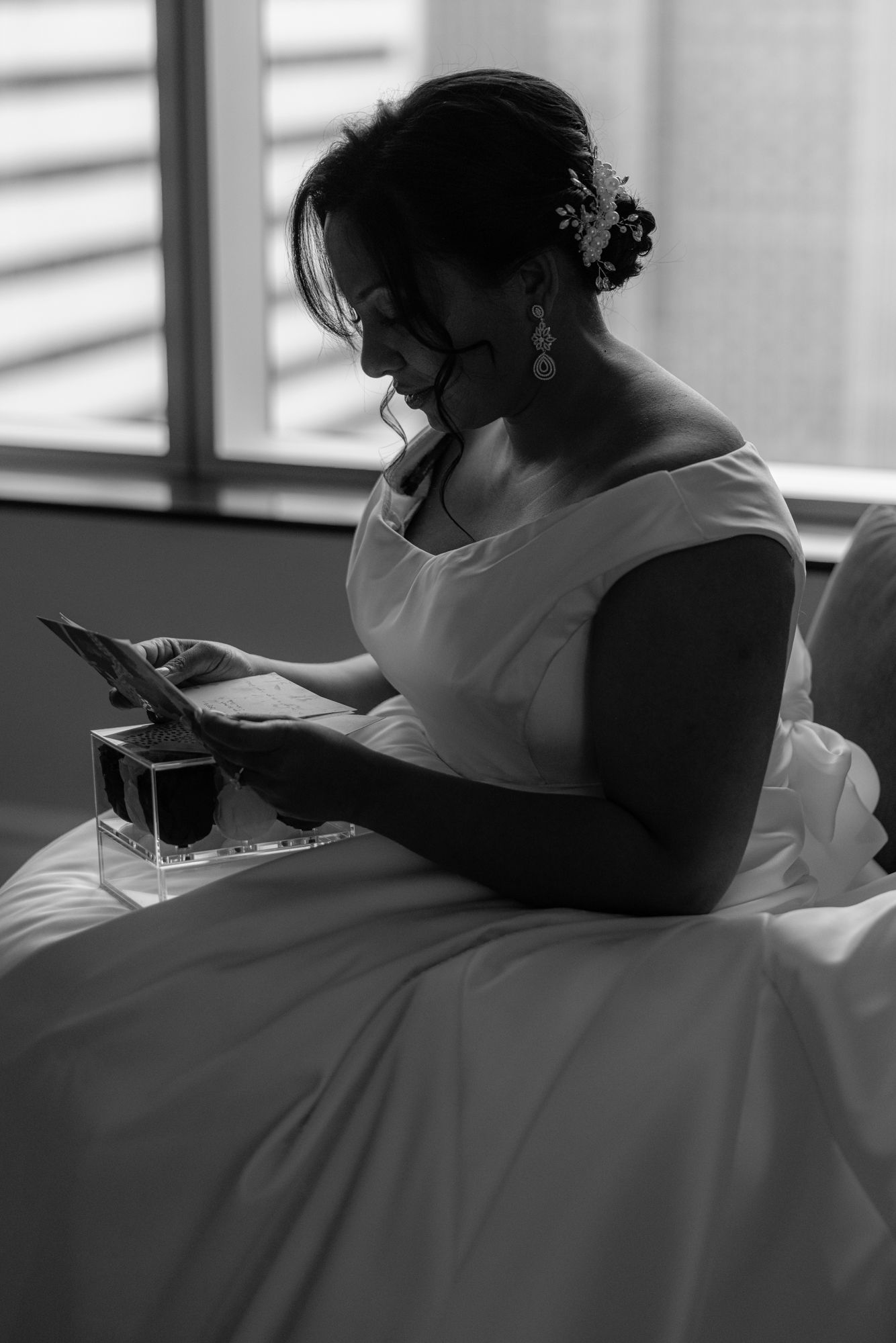 A bride in a white gown sits by a sunlit window, reading a handwritten card in a soft black and white portrait.