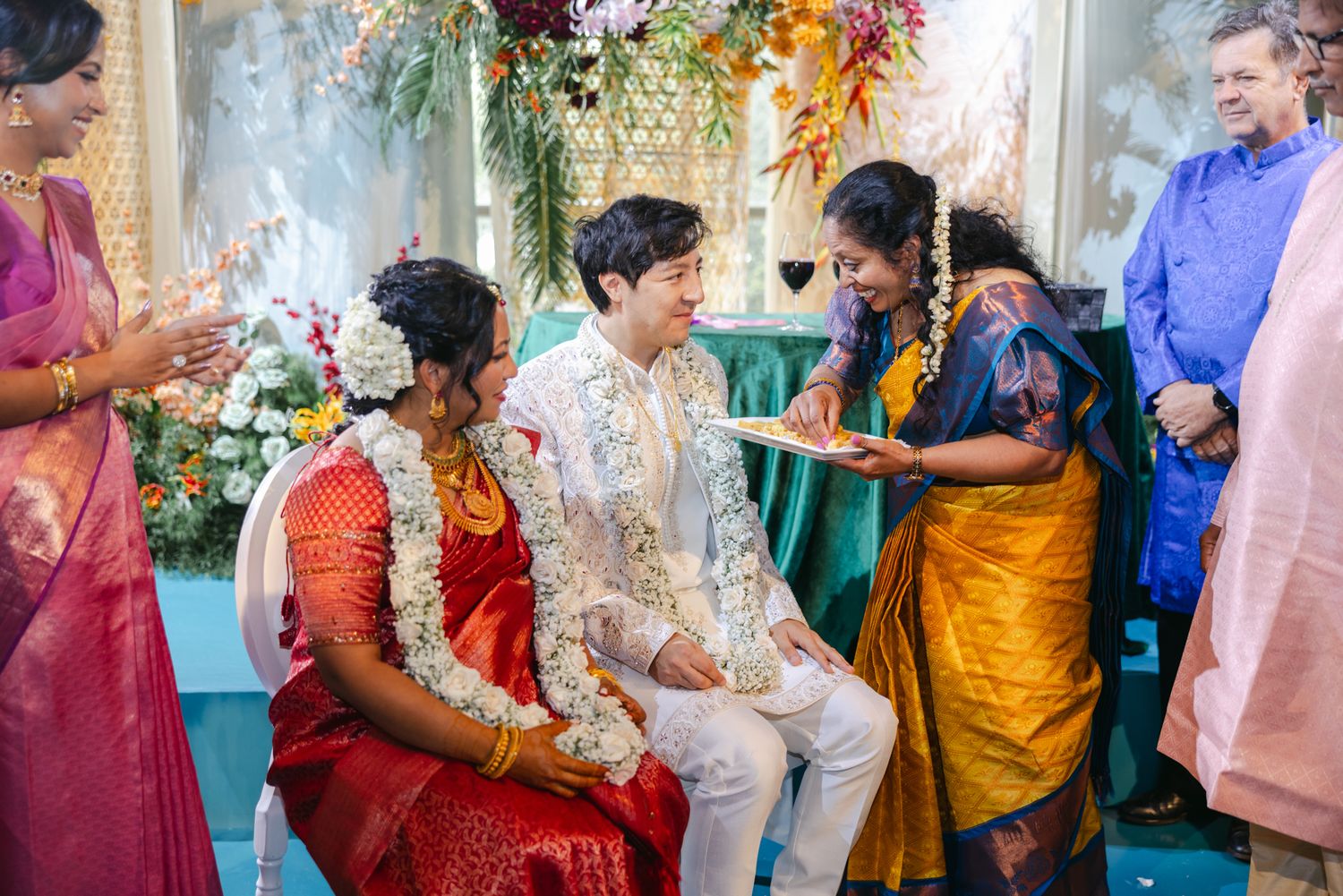 A groom and bride in a red sari wearing floral garlands sit during a traditional ceremony as relatives in colorful saris look on.