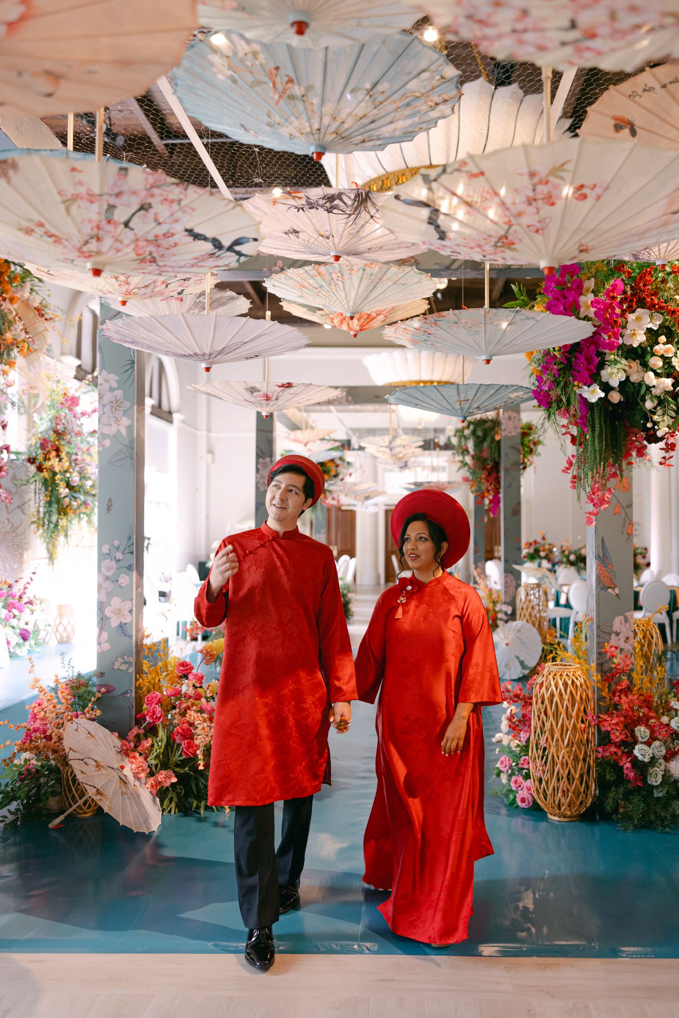 Couple in traditional red Vietnamese ao dai and headpieces walk beneath hanging floral parasols amid colorful flowers at a wedding venue.