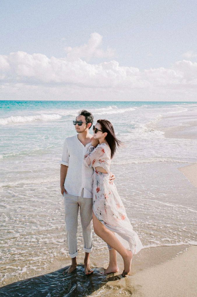 Stylish couple in sunglasses embracing on a tropical beach with turquoise waves and bright blue sky.