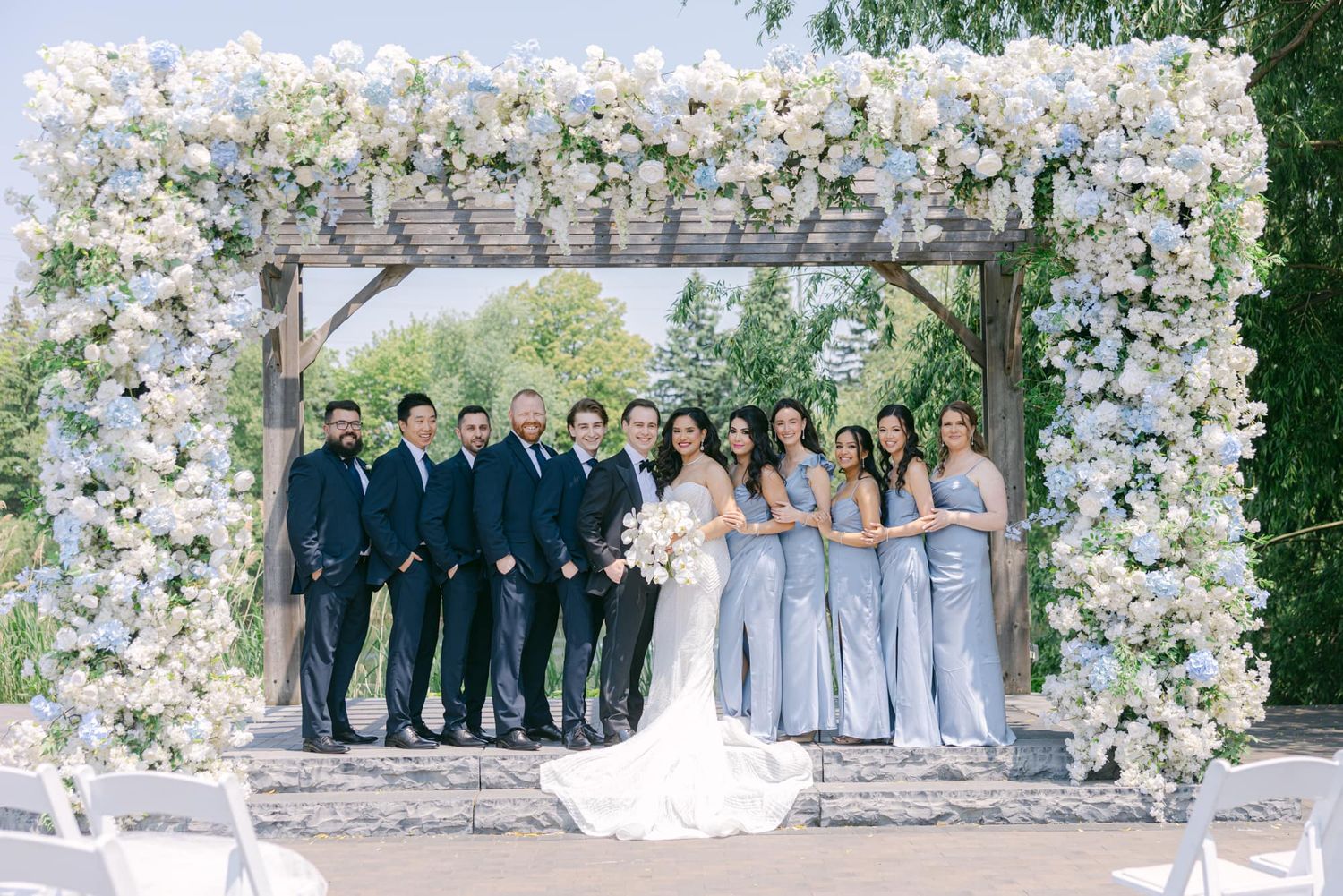 Wedding party posing under floral arch with white blooms, bridesmaids in blue and groomsmen in navy.