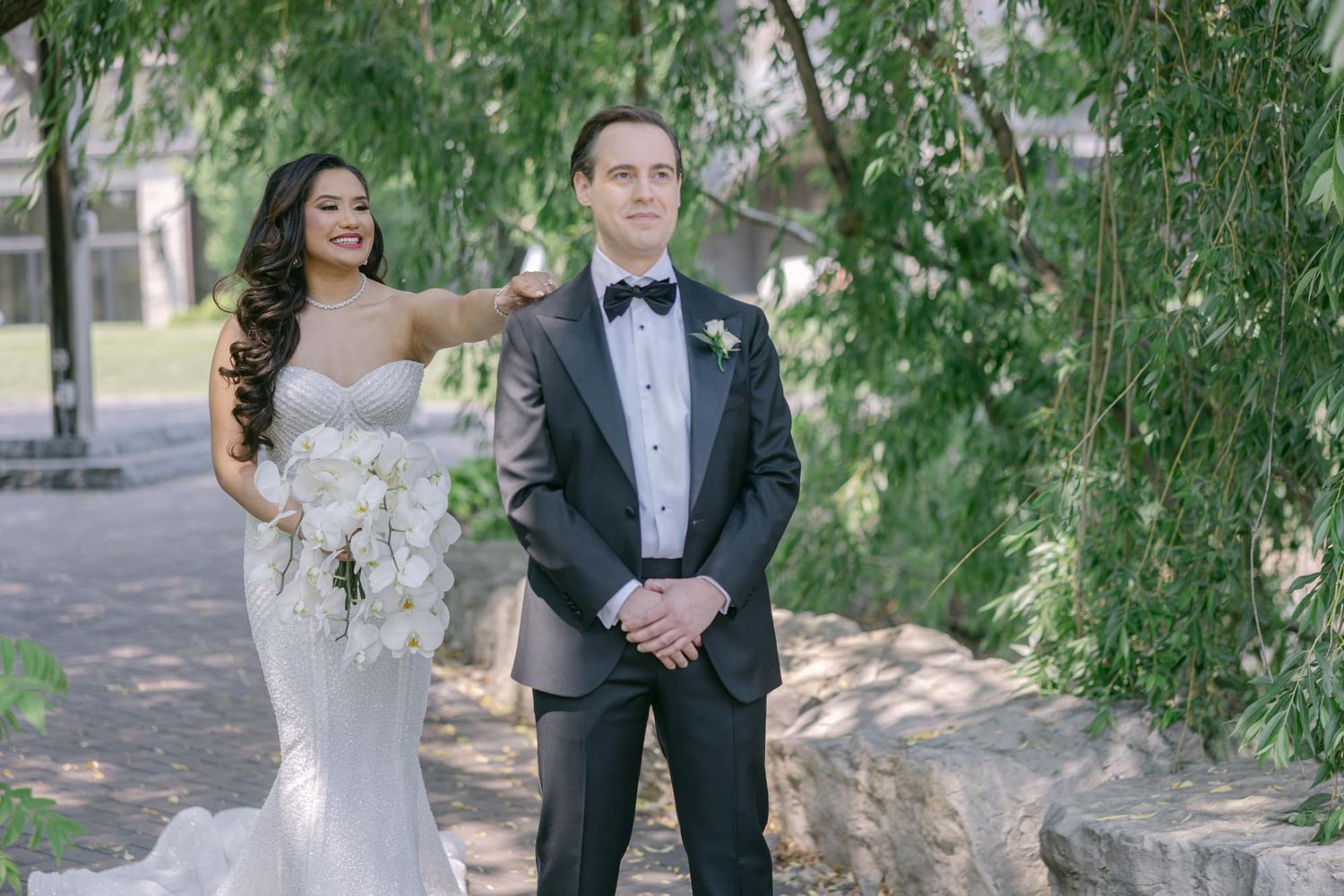Bride in a white mermaid gown tapping groom's shoulder during an emotional first look under willow trees.