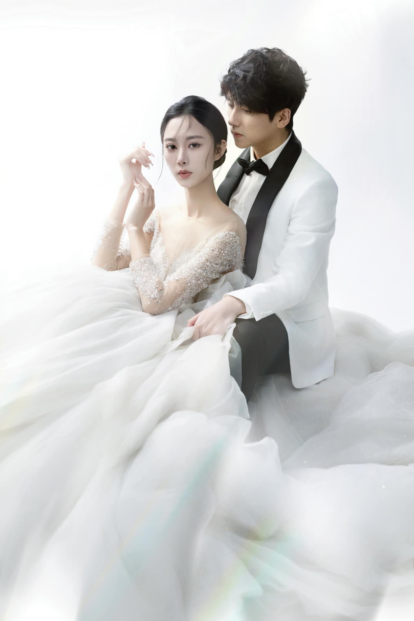 Elegant couple poses in studio wearing white wedding attire with flowing tulle gown and white tuxedo.