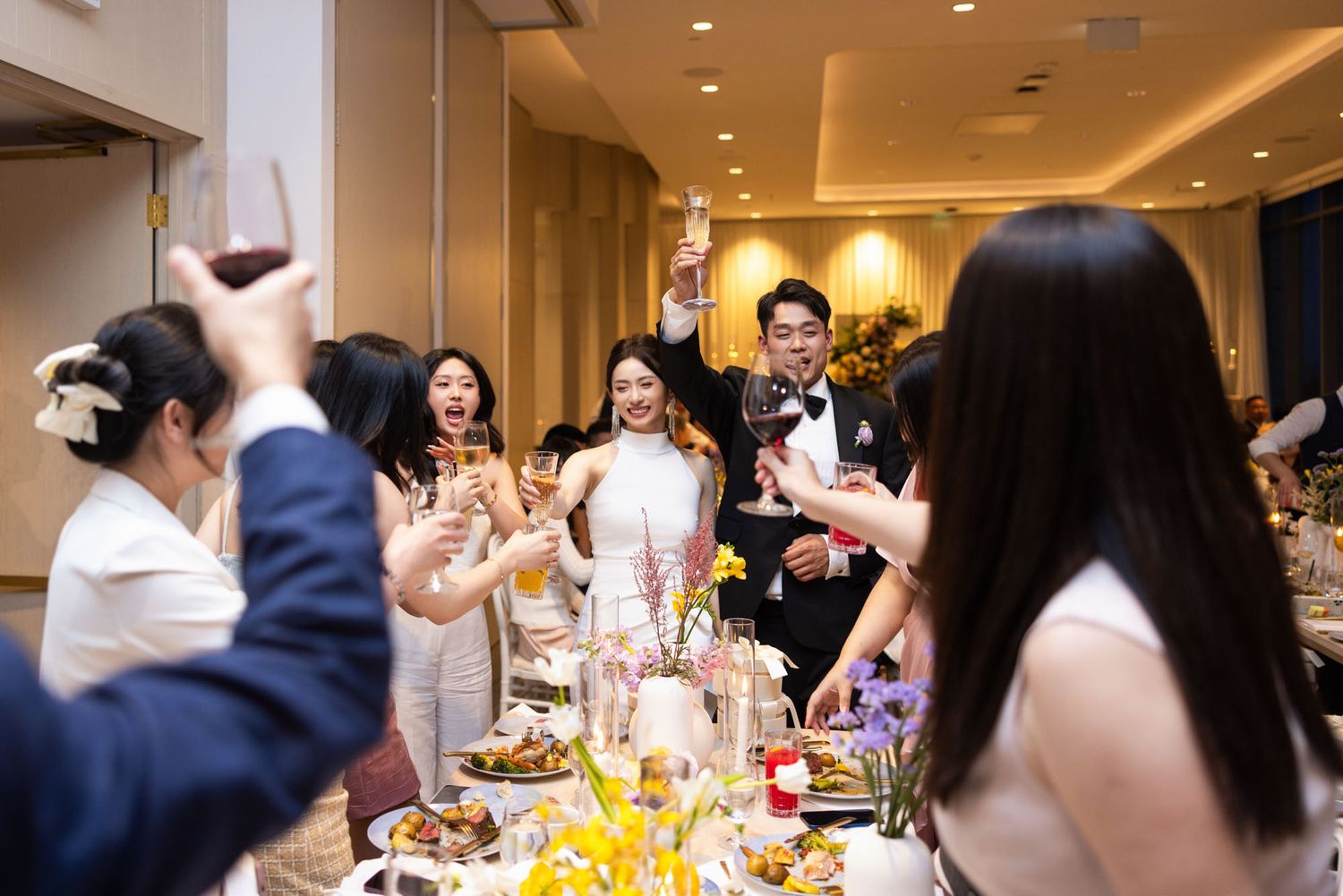 Wedding couple and guests raise glasses in a joyful champagne toast at a floral-decorated reception table.