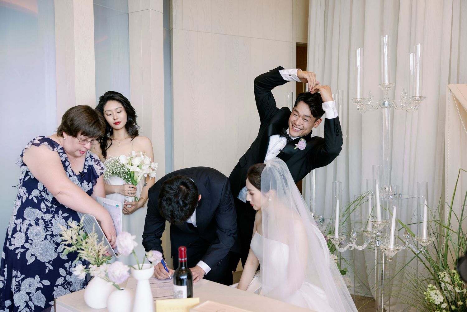 Groom strikes fun pose while wedding party signs register at elegant floral wedding ceremony.