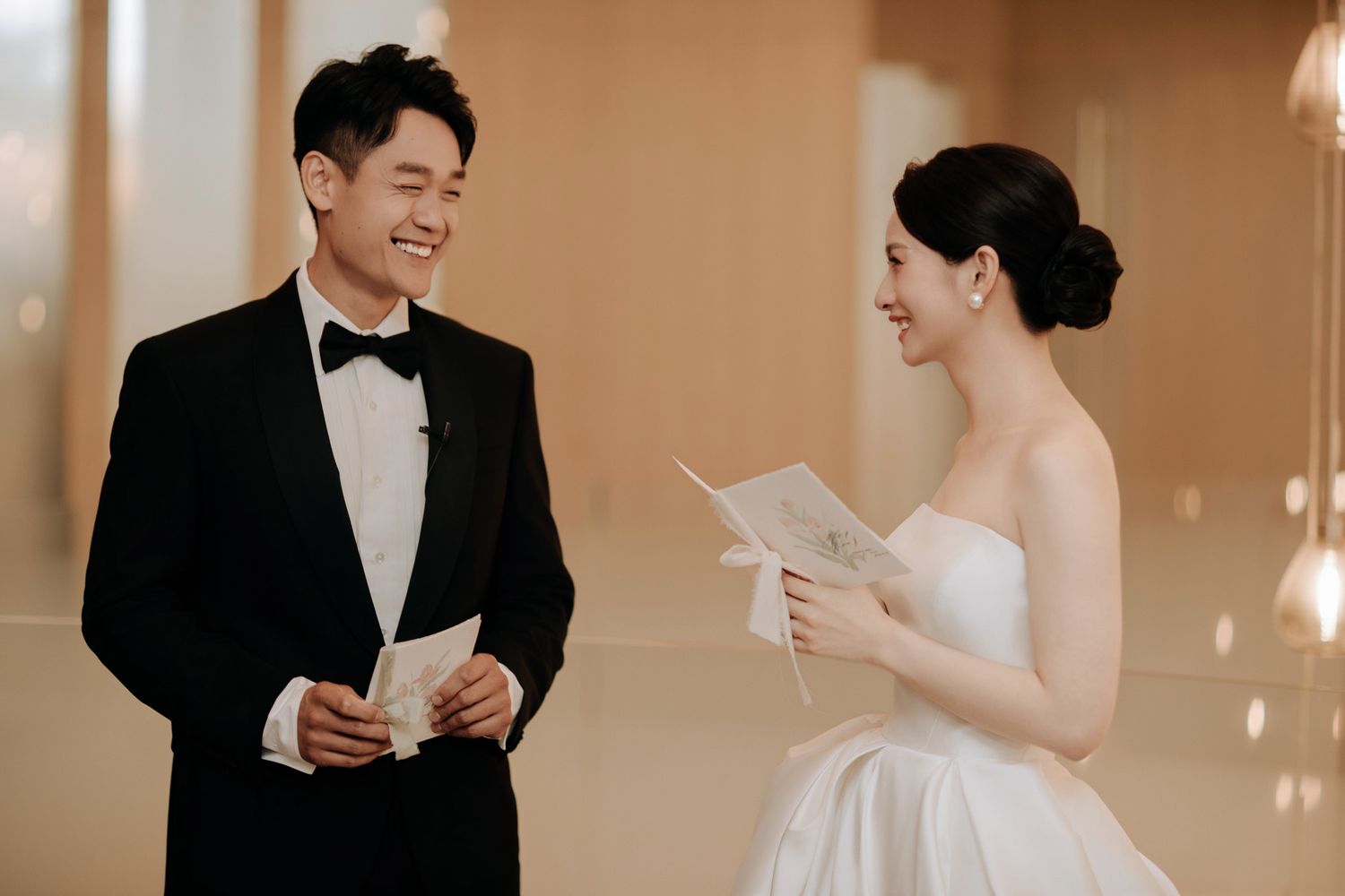 Smiling bride and groom share a joyful moment reading wedding vow letters facing each other.