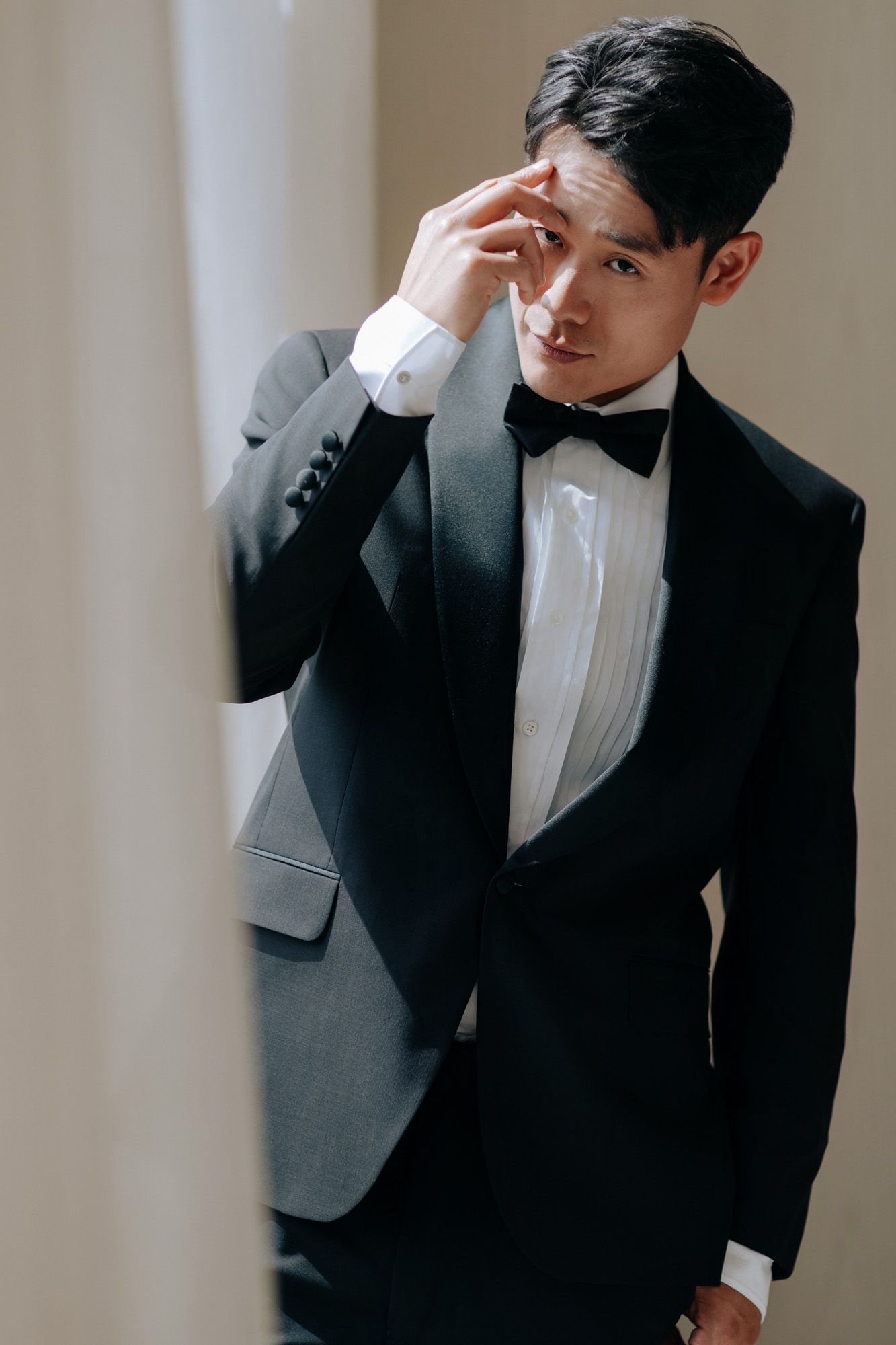 Sophisticated groom in black tuxedo adjusting hair while posing against neutral background.