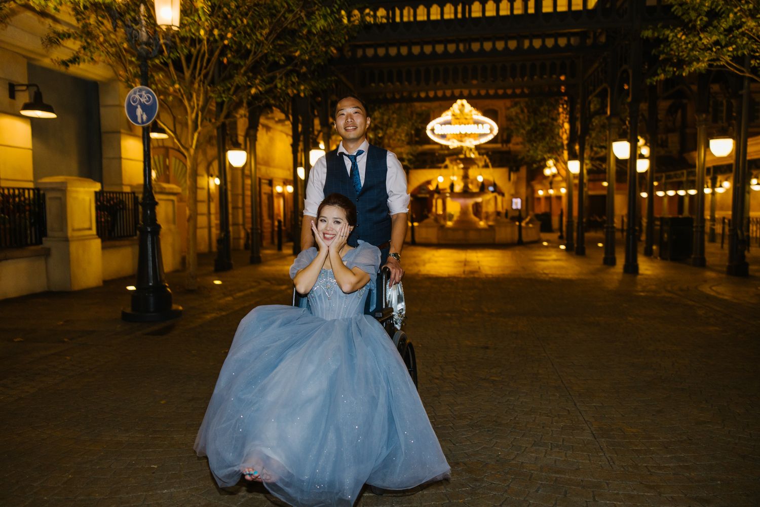 Groom pushes bride in blue gown through Disney park street at night during wedding celebration.